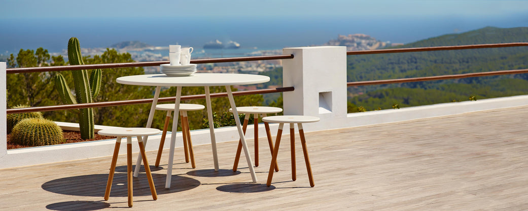 A round table with four wooden stools overlooks a scenic area featuring lush greenery and distant ocean views.