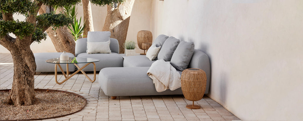 Stylish seating area with plush gray sofas, decorative pillows, a cozy throw, and natural elements like a potted tree and woven accent tables.