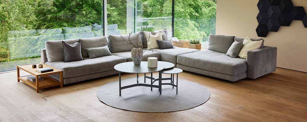 A spacious, modern living area featuring a large gray sectional sofa, a round area rug, and stylish coffee tables, with greenery visible outside.