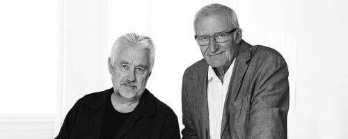 Two men posing together with friendly expressions, dressed in casual attire against a light background, conveying collaboration and creativity.