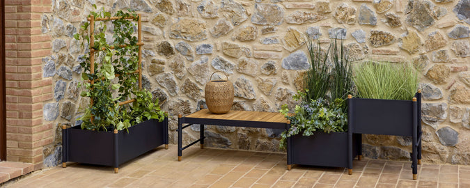 A display of stylish planters with various green plants, complemented by a wooden bench against a rustic stone wall.