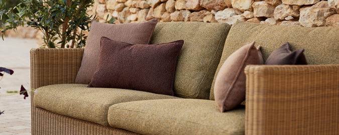 A cozy seating area with textured green cushions and decorative pillows in shades of purple and brown against a stone backdrop.