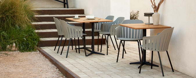 A stylish cafe setting featuring wooden tables and grey chairs arranged on a paved area, complemented by greenery in the background.