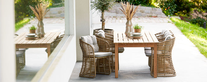 A stylish wooden table surrounded by woven chairs, with decorative plants and greenery in the background, perfect for gatherings.