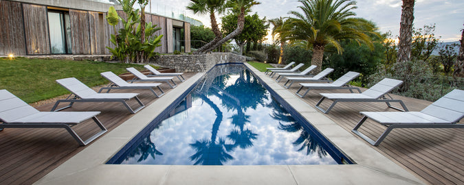 A serene poolside scene featuring a row of white sunbeds beside a tranquil water, surrounded by lush greenery and palm trees.