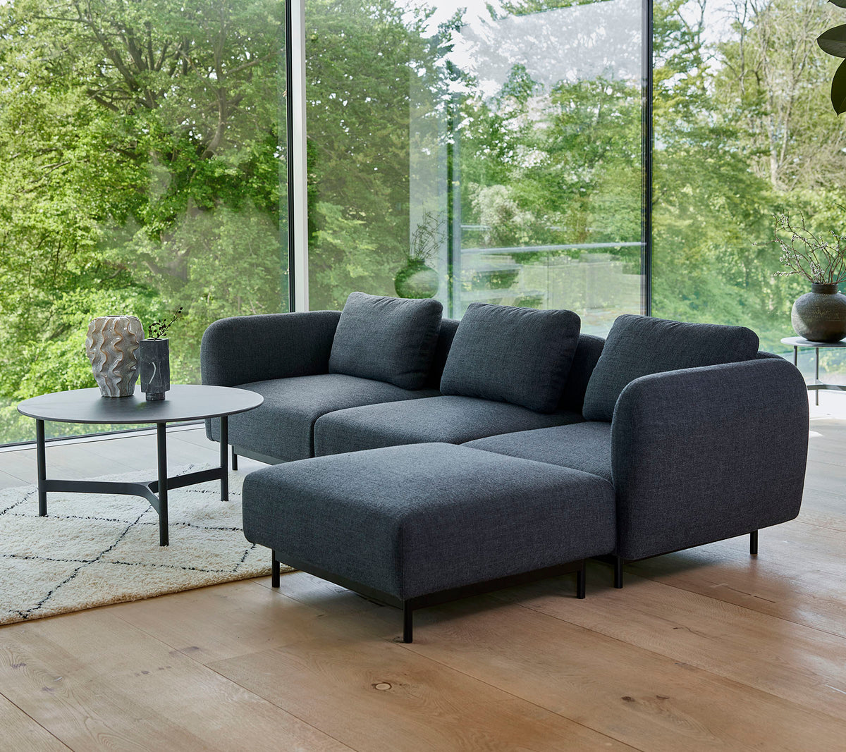 A modern gray sectional sofa with plush cushions, paired with a sleek round coffee table on a light rug, surrounded by greenery.