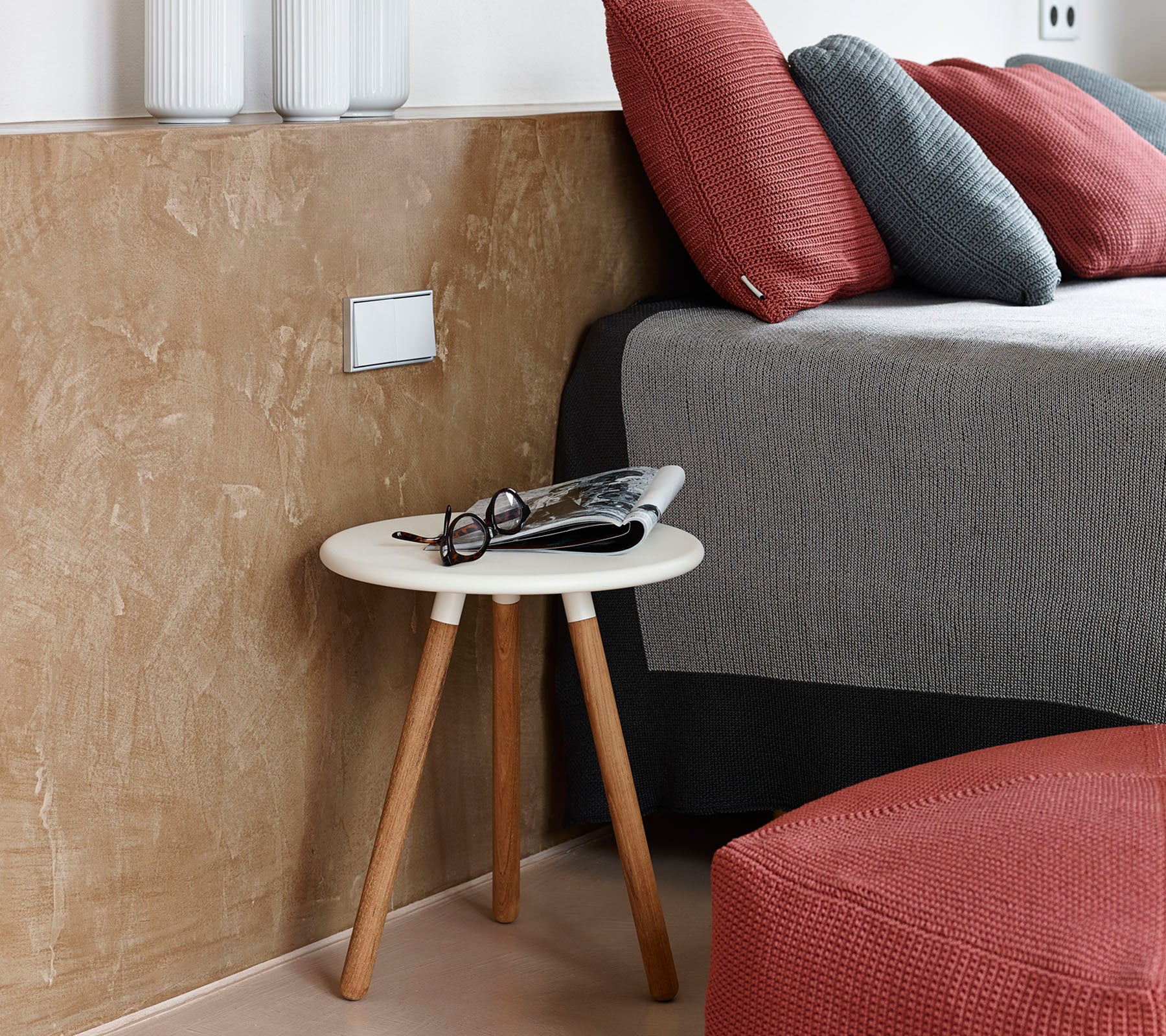 A cozy bedroom scene featuring a low side table with glasses and a magazine beside a neatly made bed with textured pillows.