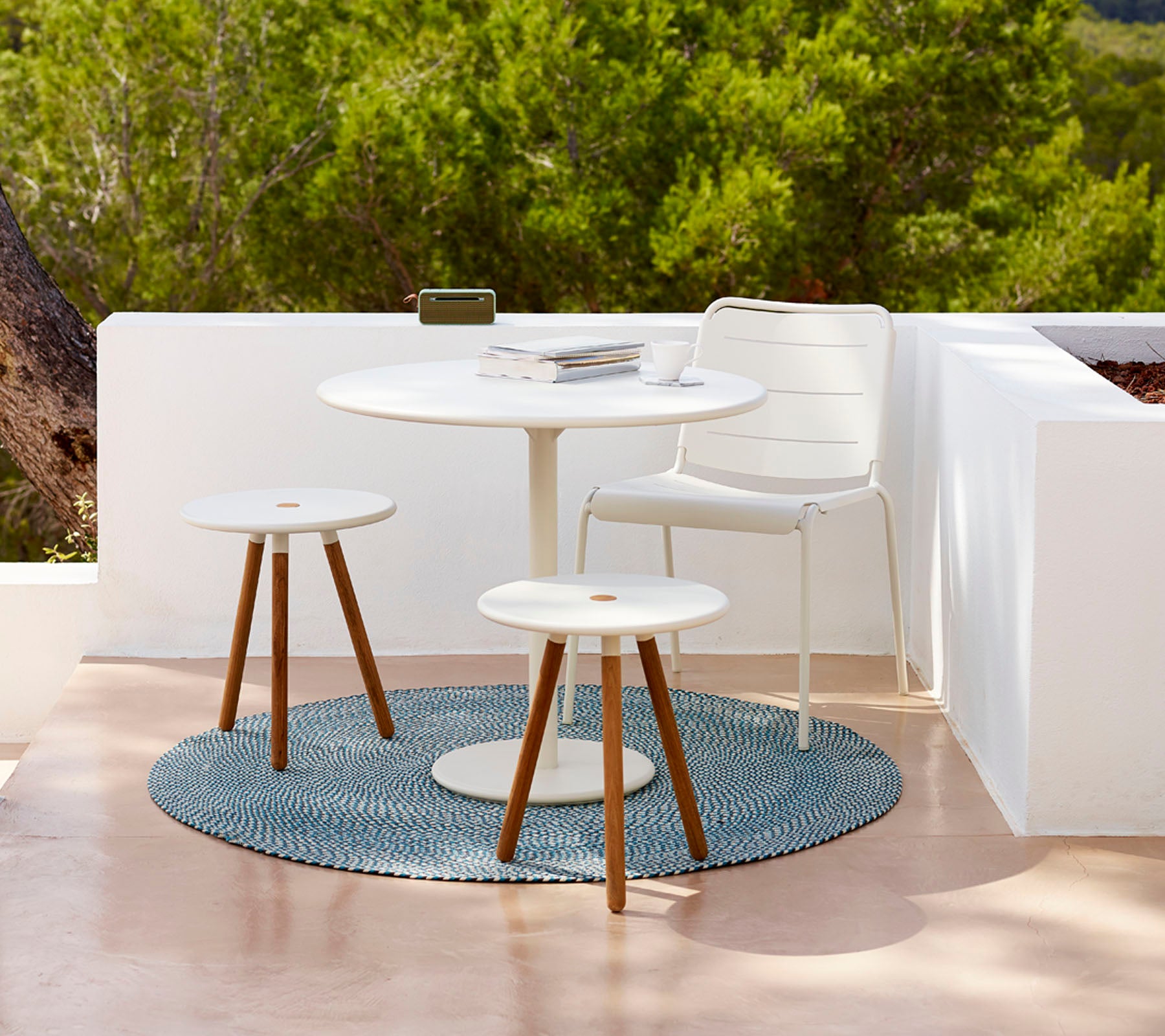 Modern outdoor table set featuring a round table, two stools, and a textured rug, surrounded by greenery. Ideal for relaxation.