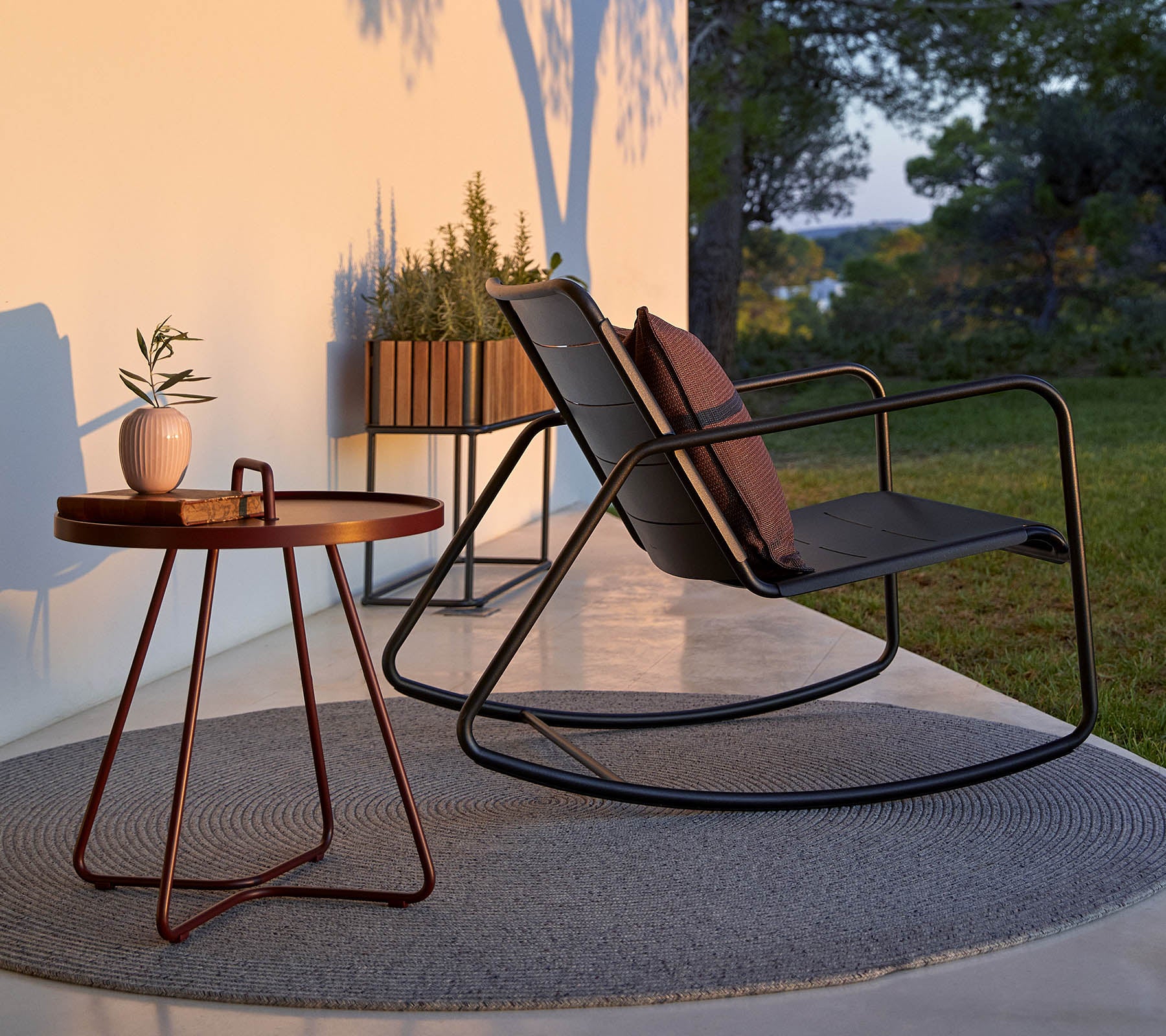 A stylish rocking chair with cushions, accompanied by a small round table and a potted plant, set against a serene backdrop.