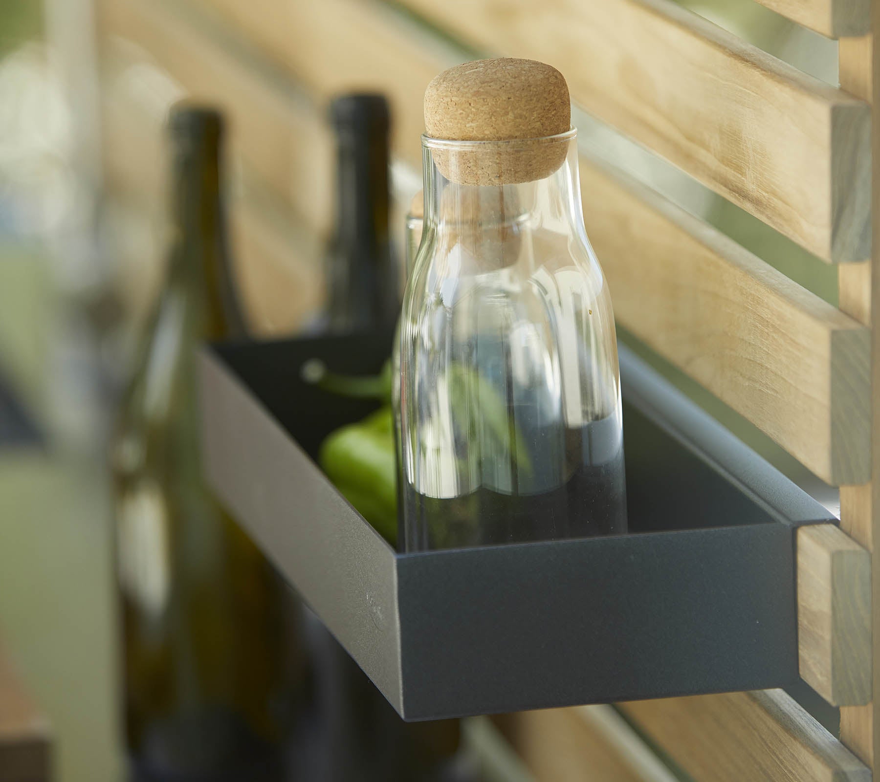 A glass bottle with a cork stopper sits on a shelf alongside several dark bottles, set against a wooden backdrop.