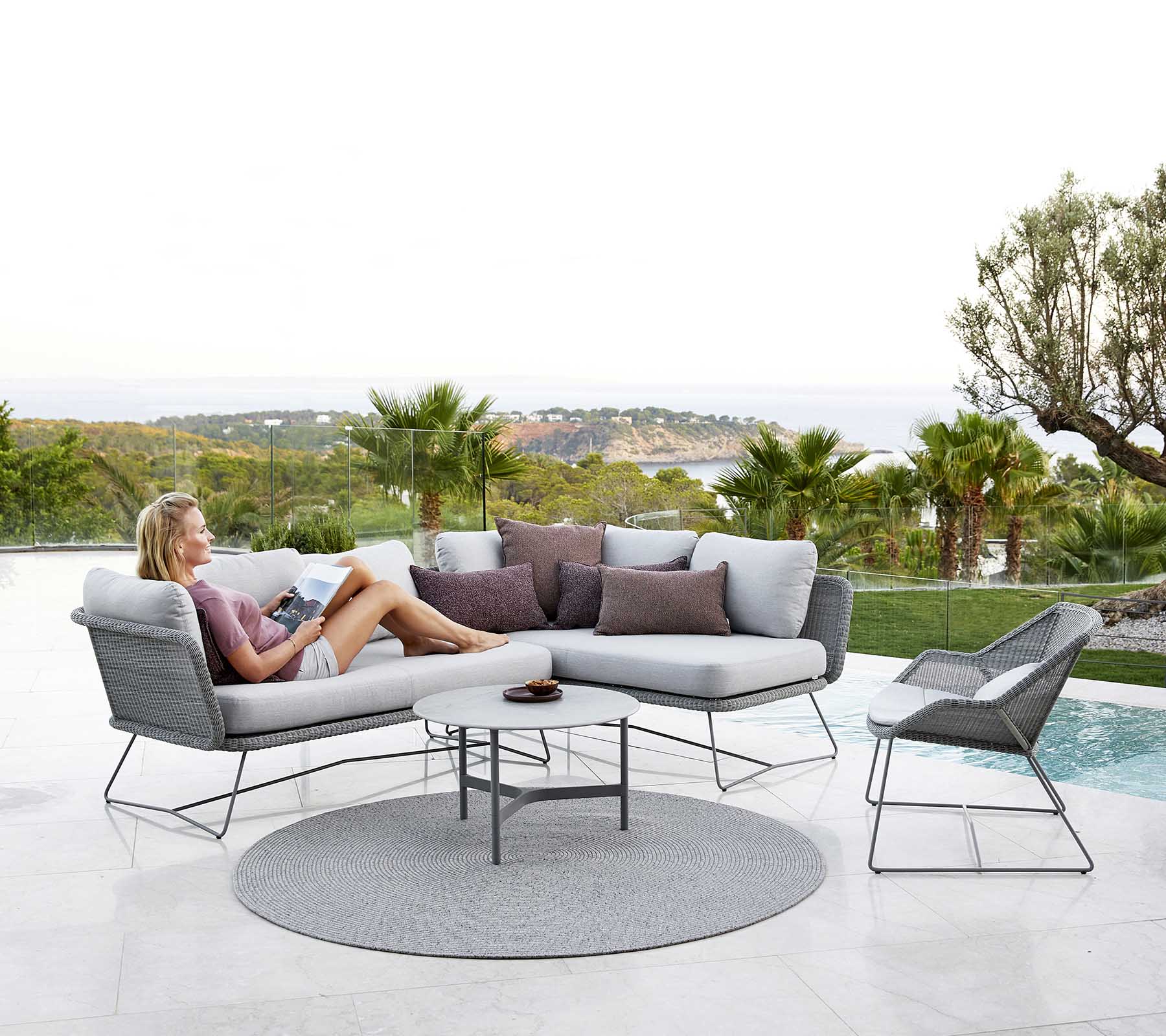 A modern outdoor seating arrangement featuring a woman reading, with cushions and a circular rug beside a pool and palm trees in the background.