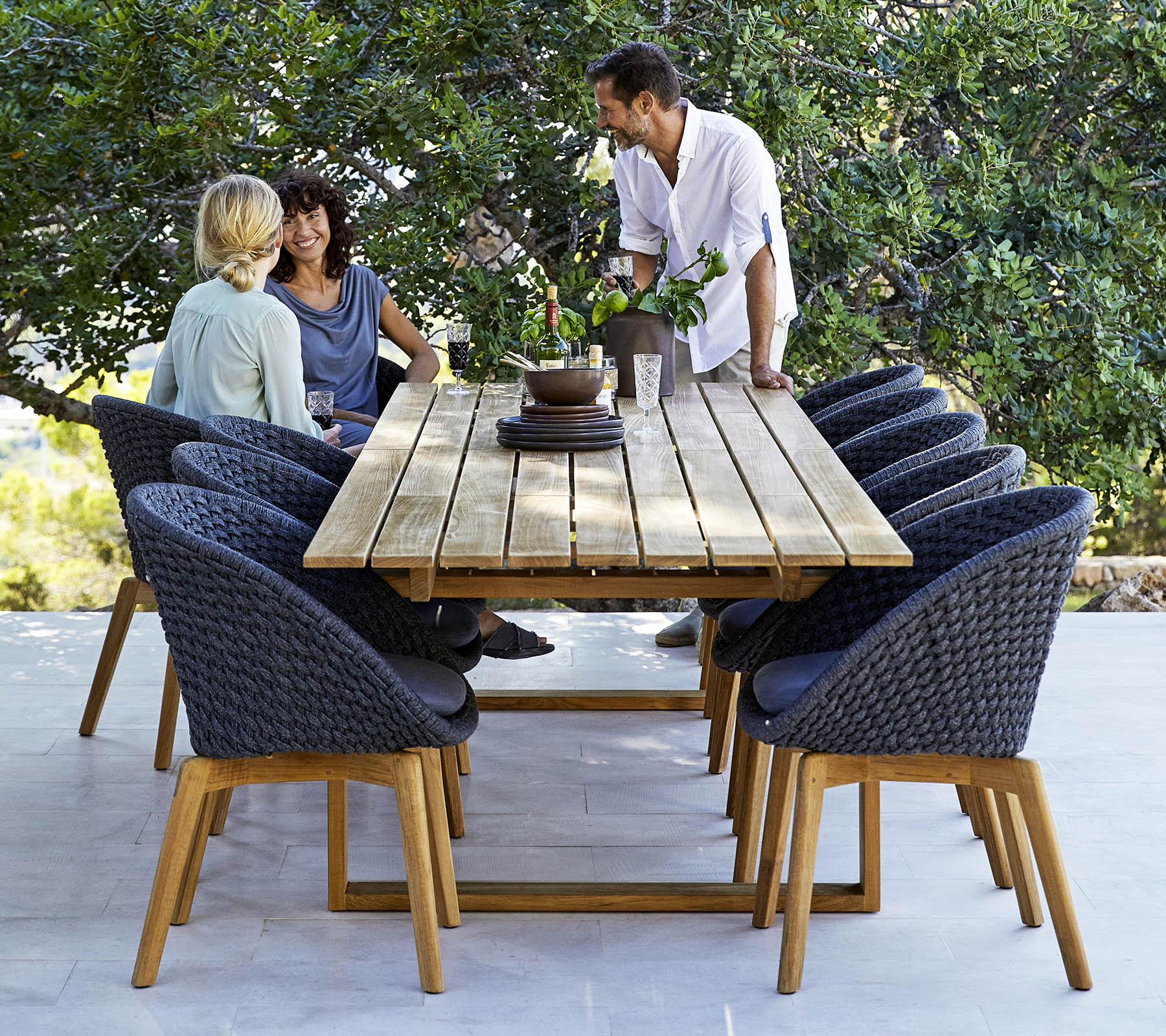 A stylish dining setup featuring a wooden table surrounded by comfortable chairs, with three people engaged in conversation, amidst greenery.