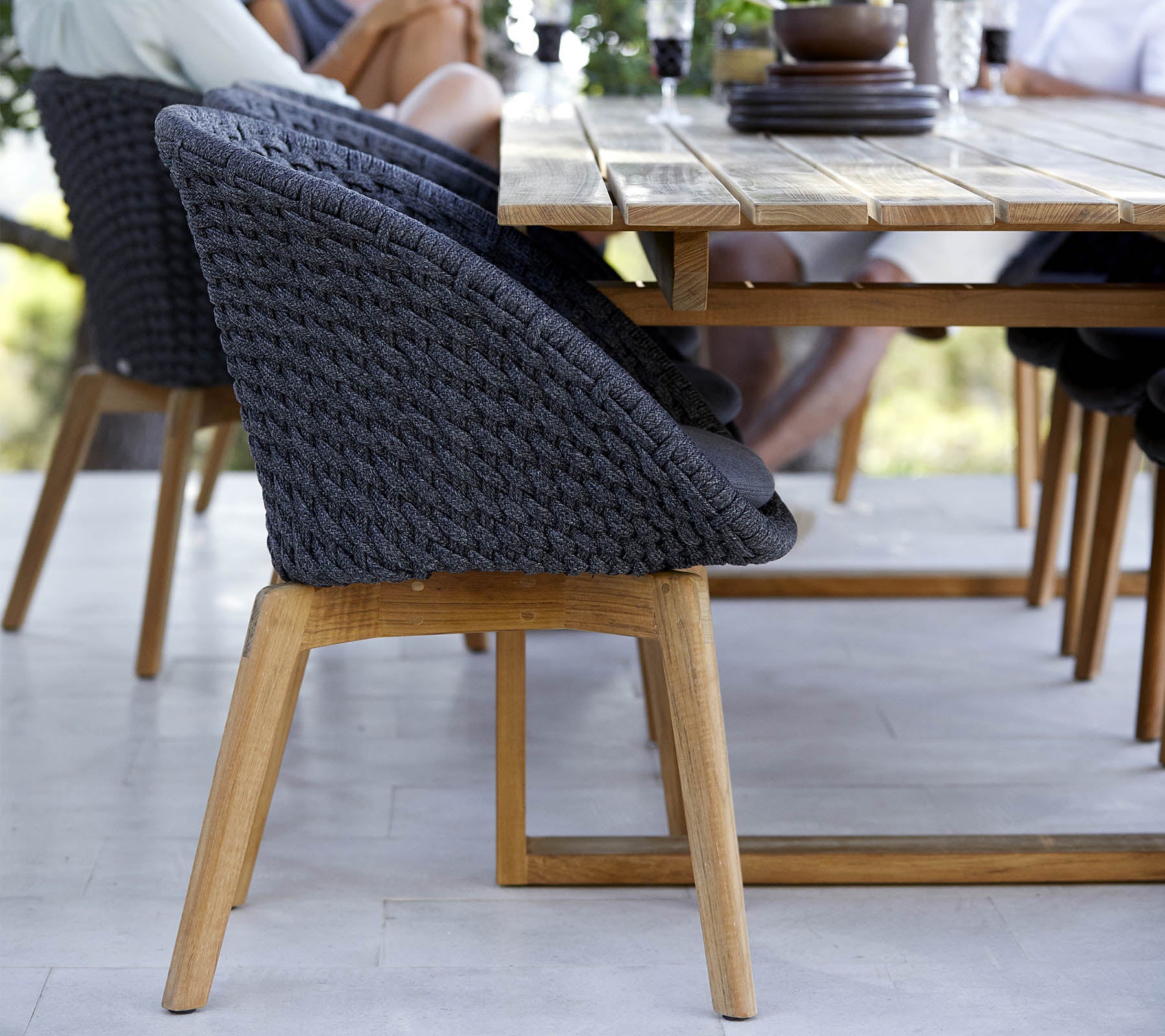 A stylish chair with a woven back and wooden legs, positioned beside a wooden dining table, with people engaged nearby.