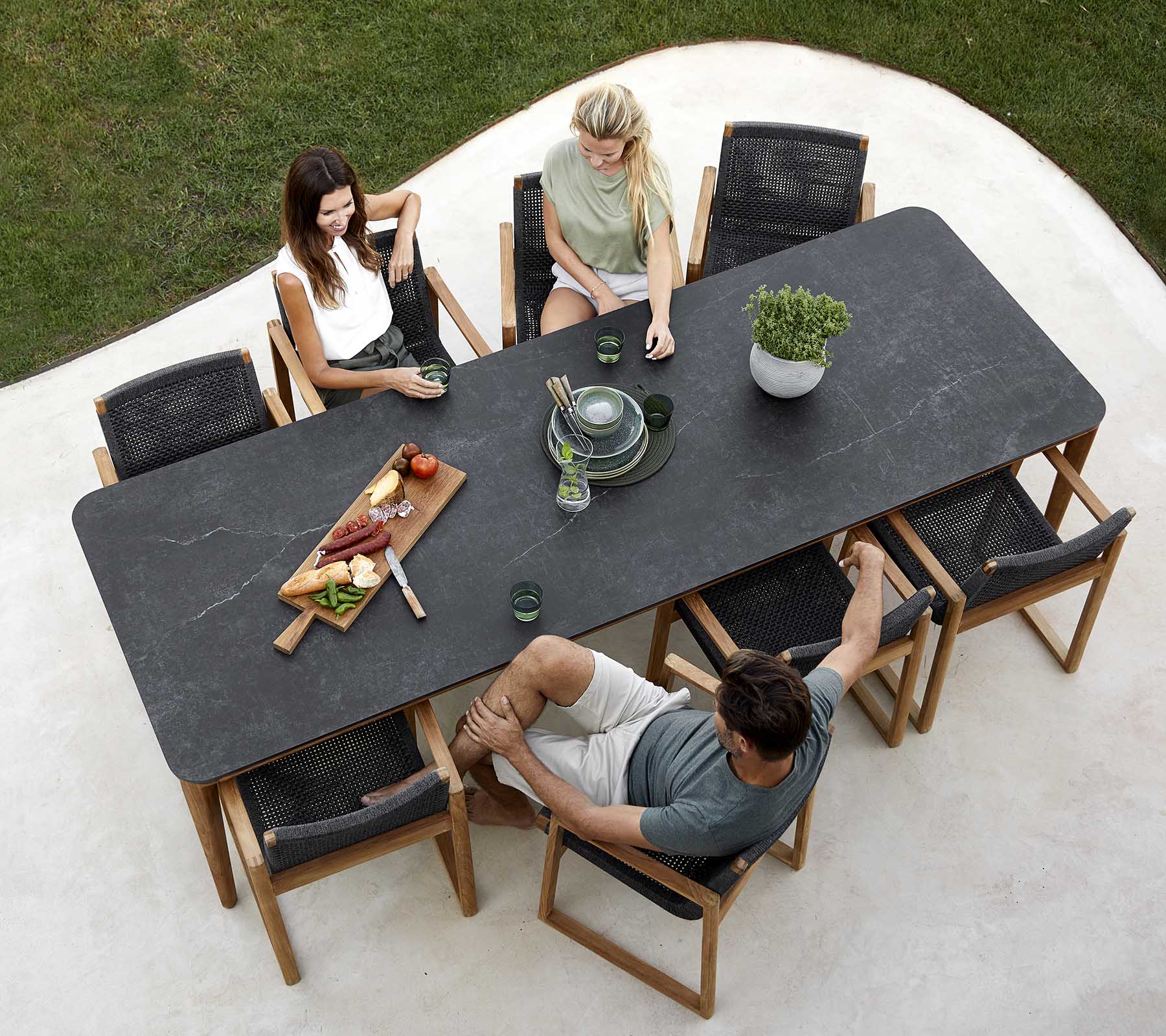 A group of people enjoying a meal around a large table, with dishes, drinks, and a plant centerpiece. Casual dining atmosphere.