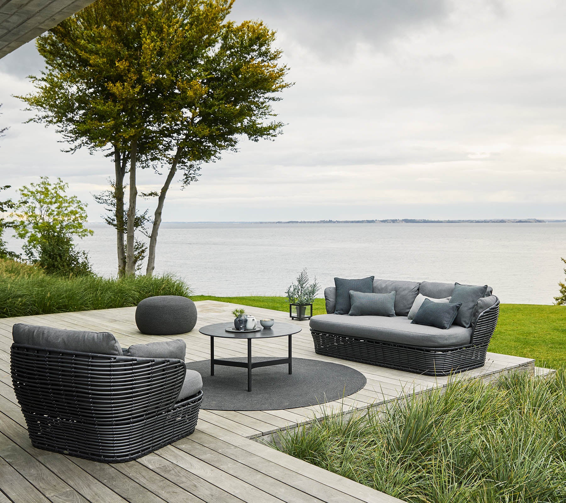Cozy seating arrangement with a sofa and chairs set around a circular table, surrounded by greenery and a water view.