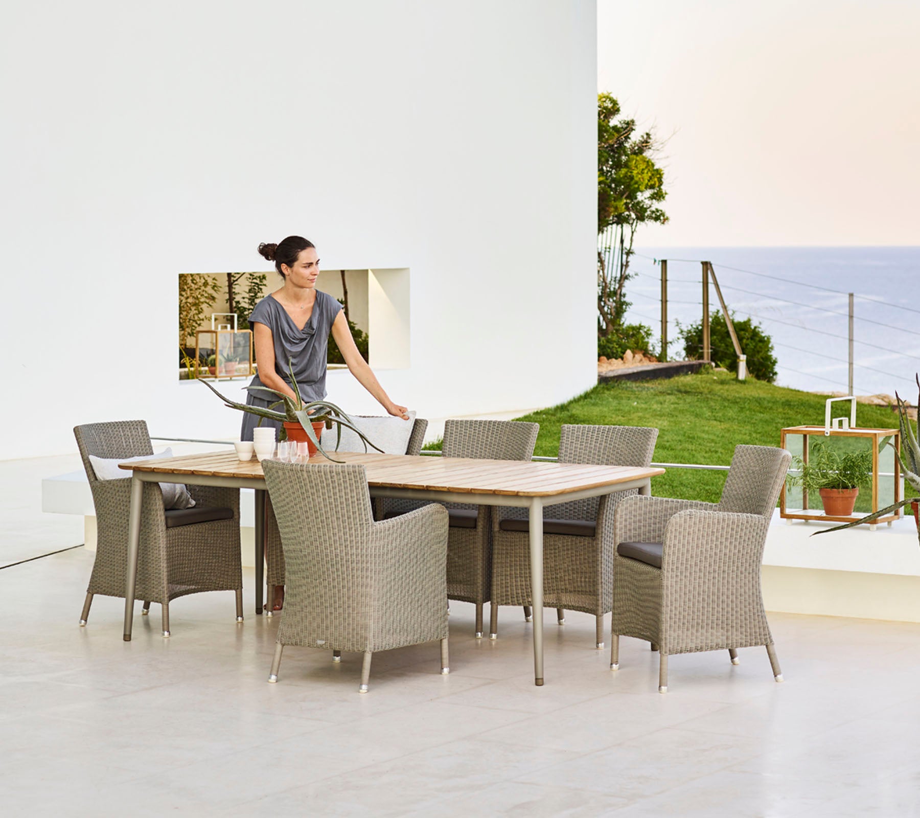A woman sets a dining table surrounded by wicker chairs, overlooking a scenic view with greenery and water in the background.