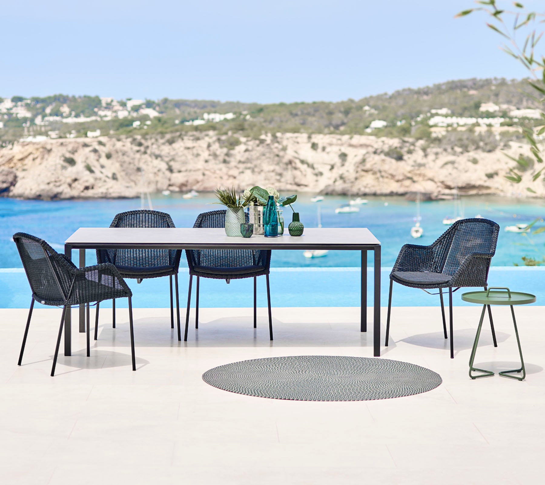 A modern outdoor dining set featuring a rectangular table, woven chairs, and a small side table, with a scenic beach backdrop.