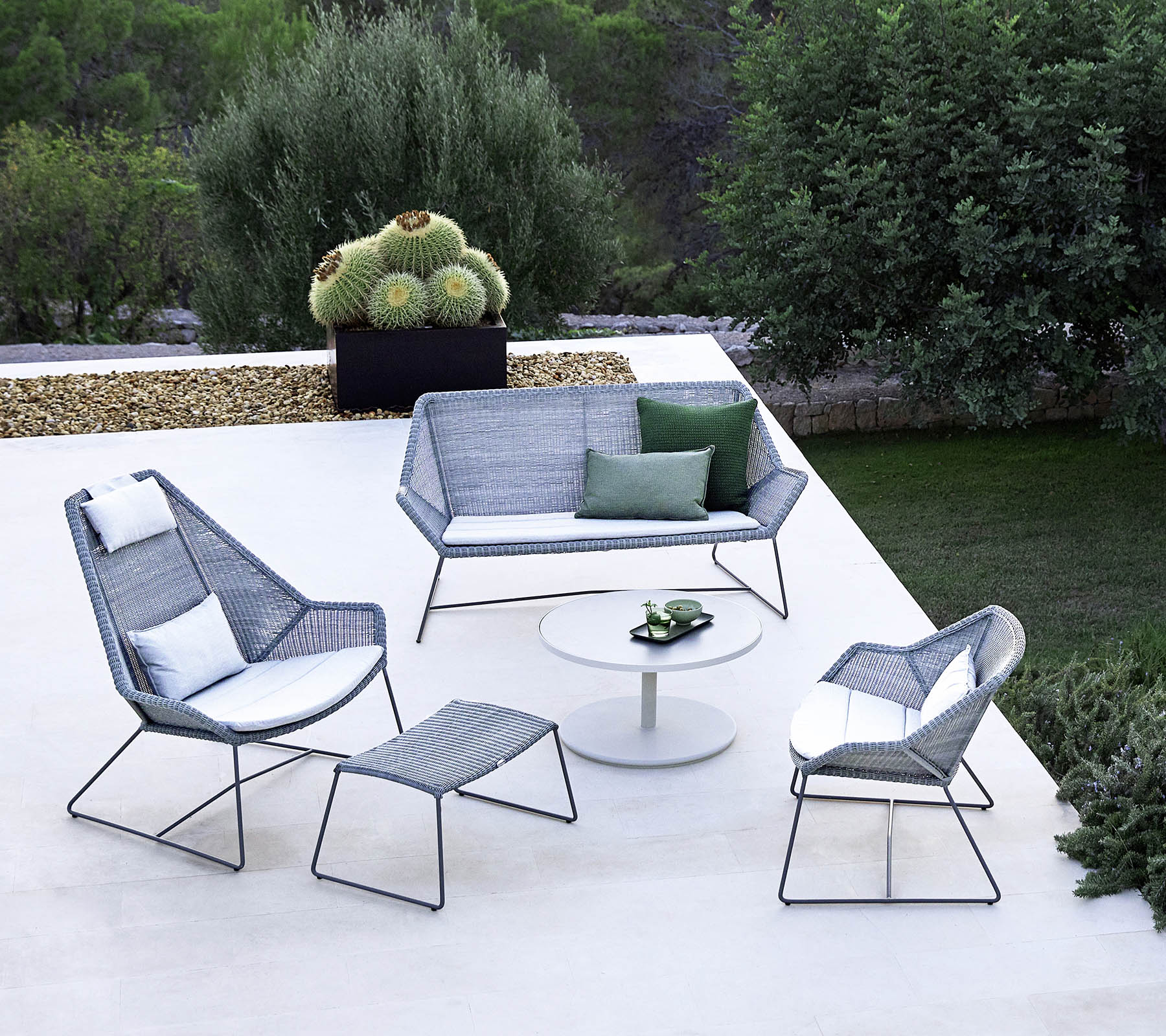 Stylish outdoor seating arrangement featuring a sofa, two armchairs, a footrest, and a coffee table, complemented by greenery in the background.