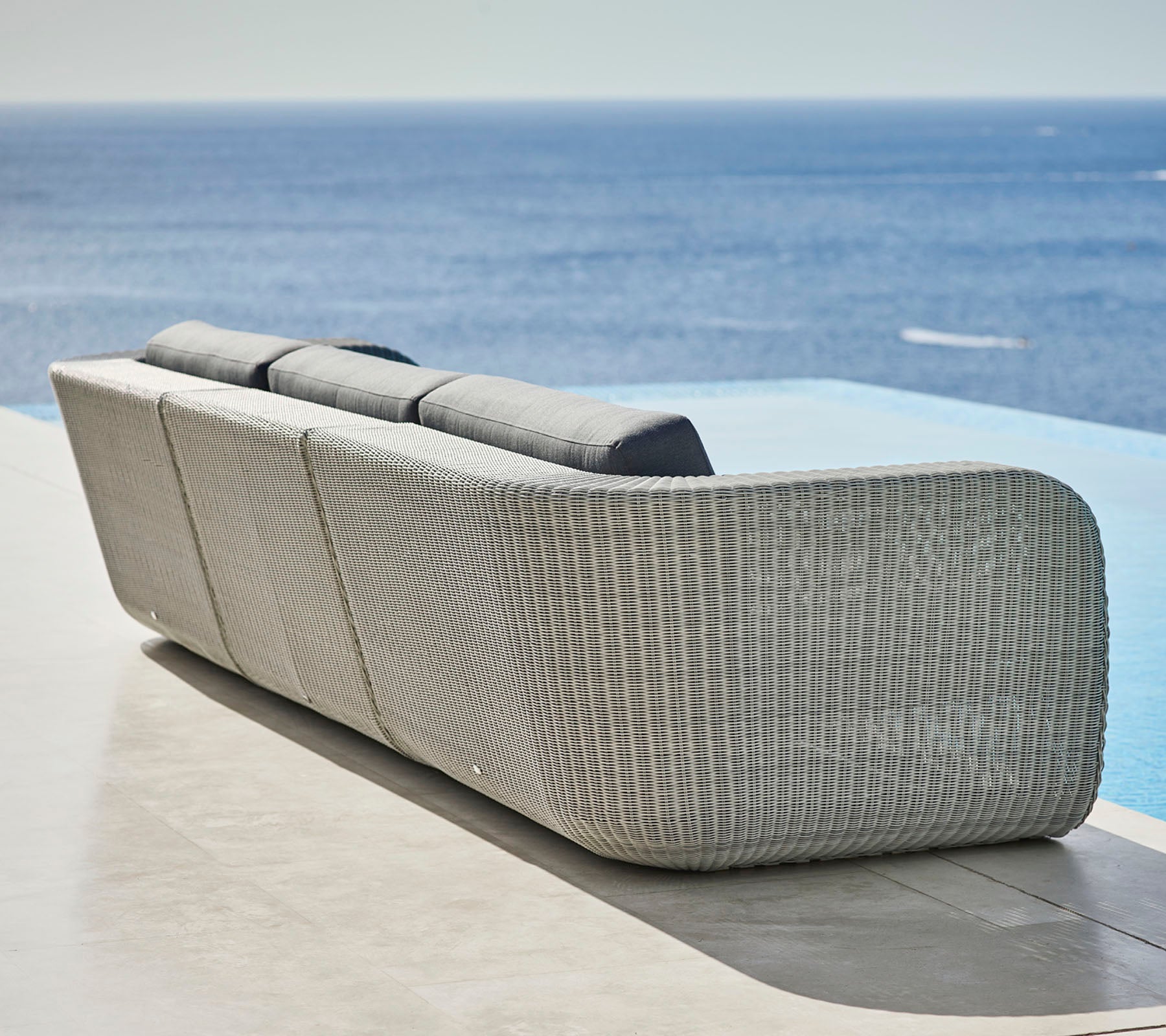 Stylish woven sofa with curved design and plush cushions, overlooking a tranquil ocean view. Ideal for relaxation and outdoor settings.