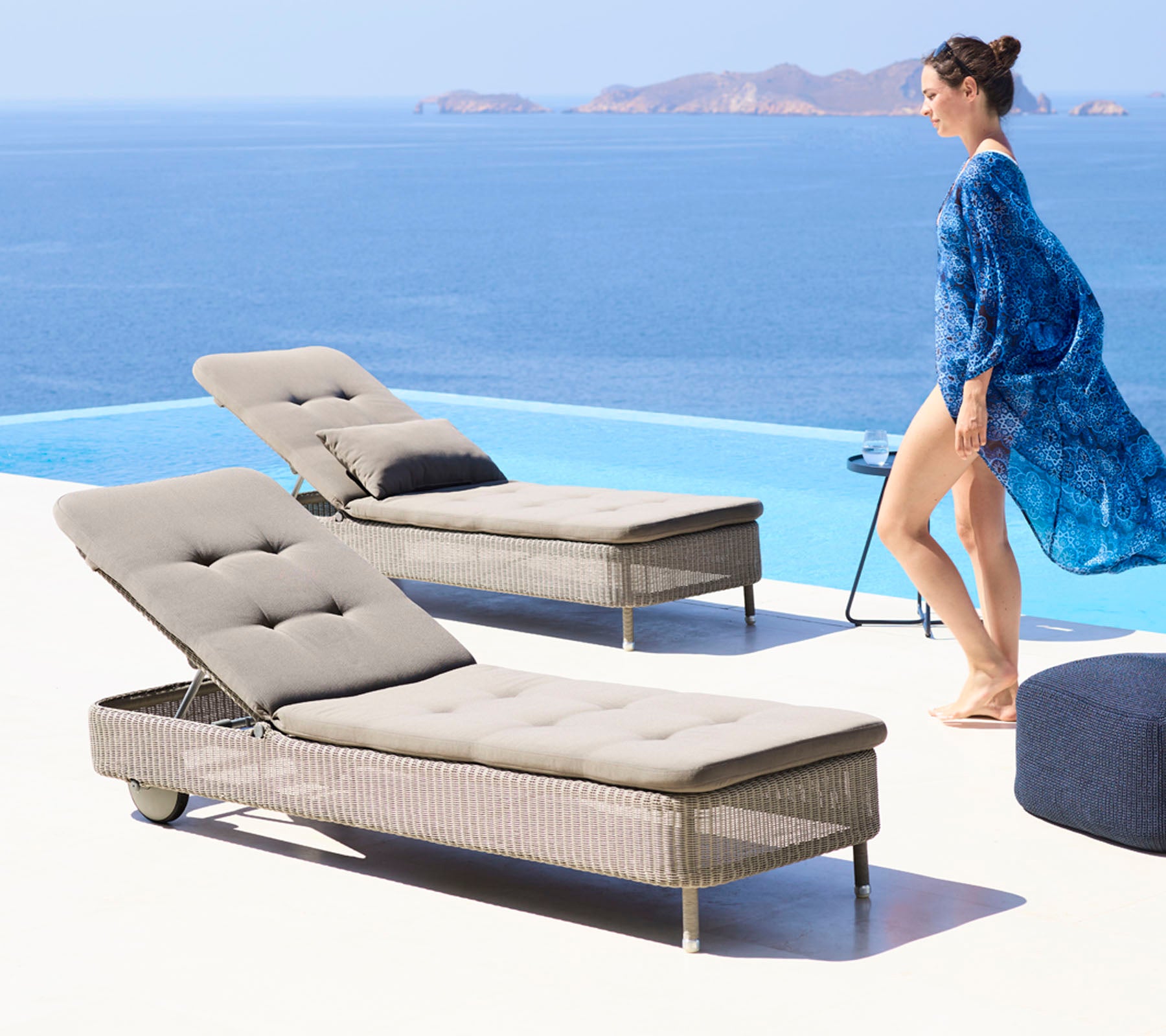 A woman in a flowing blue cover-up walks beside two stylish lounge chairs by a pool, overlooking a serene ocean view.