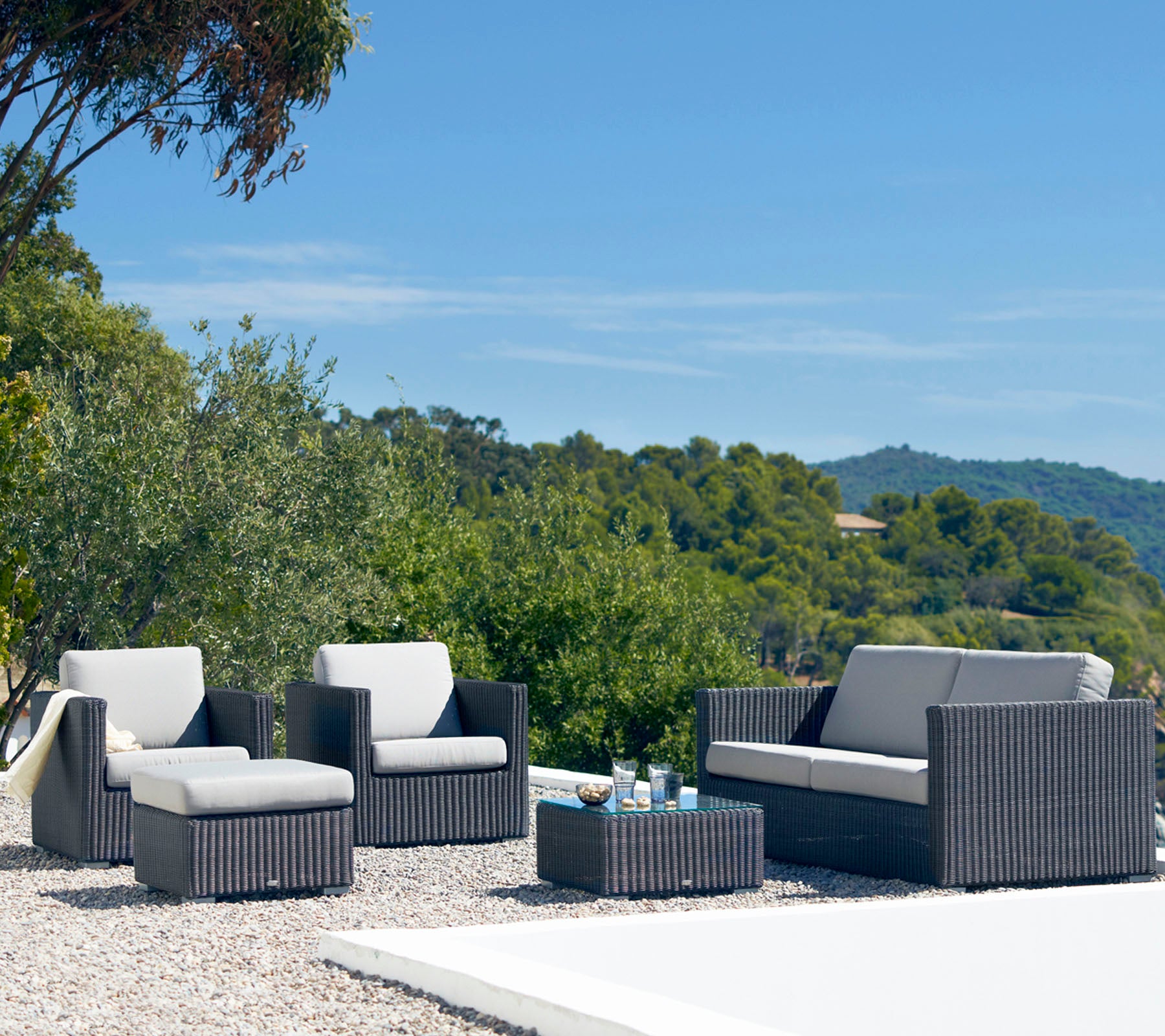 Stylish outdoor seating set featuring a sofa, two armchairs, and a coffee table with a sleek design and neutral cushions surrounded by greenery.