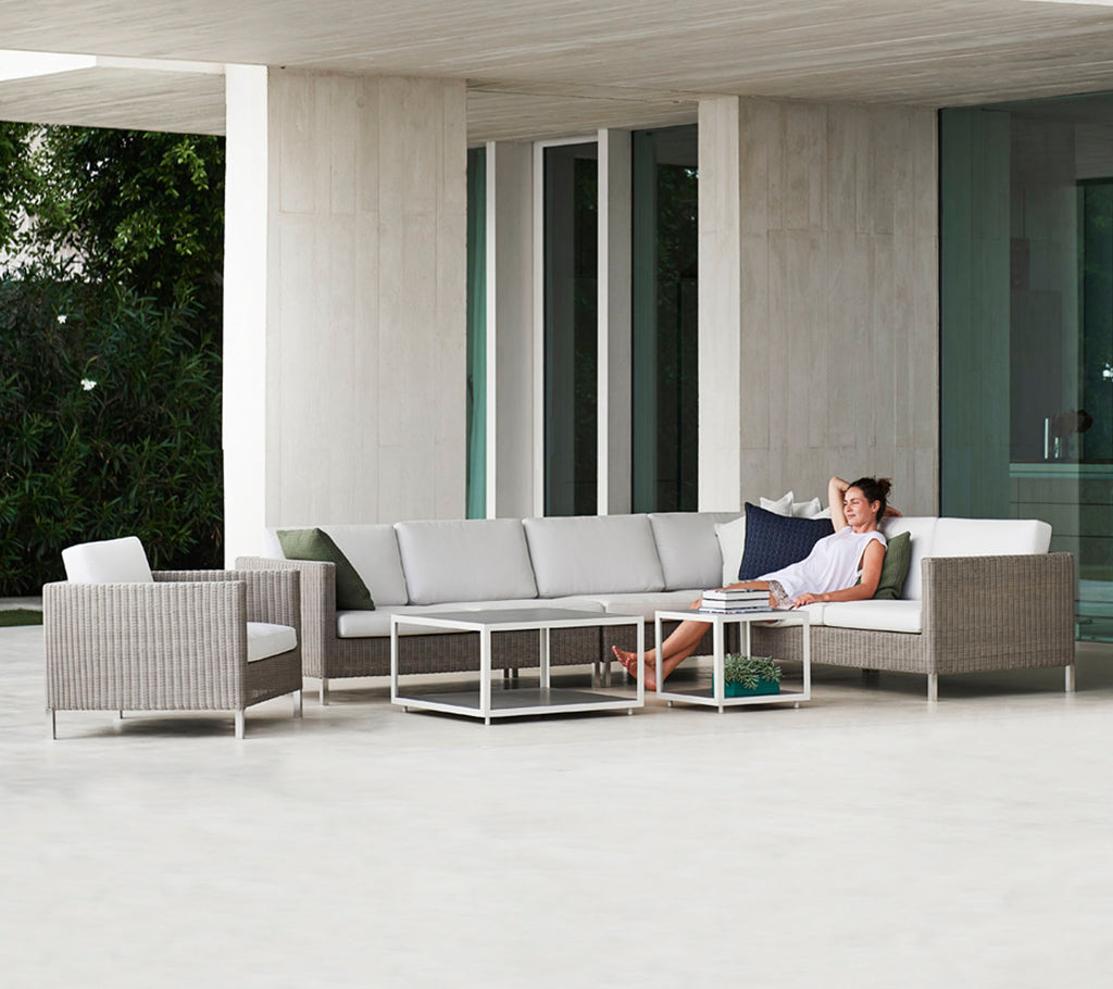 A modern seating arrangement with a gray sectional sofa, a glass-top table, and a wicker chair, featuring a person relaxing comfortably.