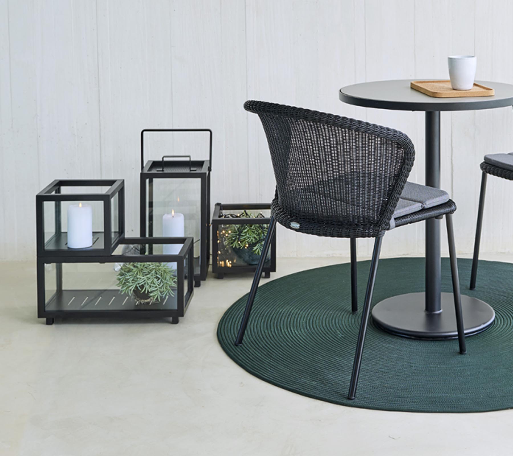 Modern seating and decorative glass lanterns with plants, arranged around a round table, on a round green rug.