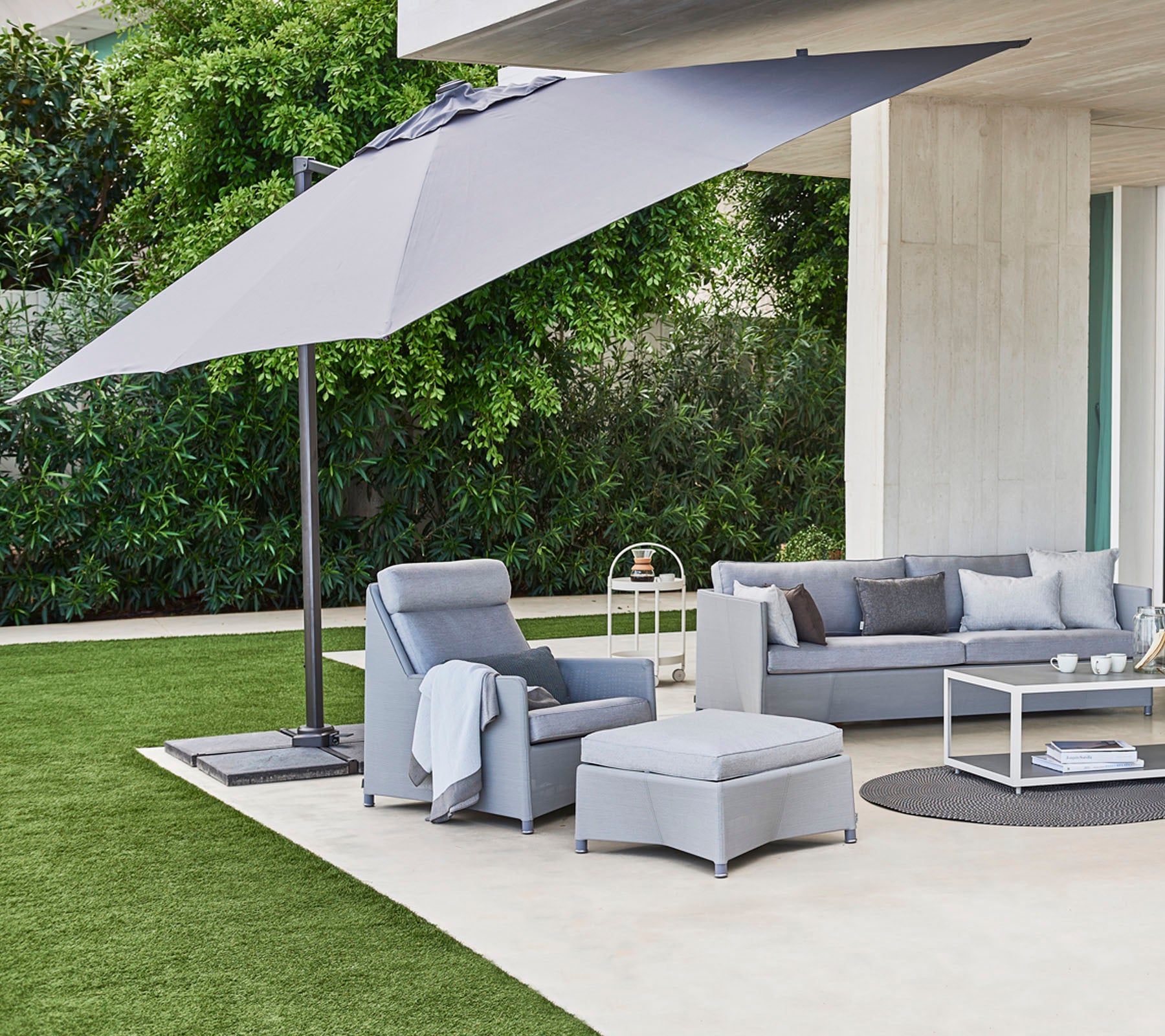 Stylish outdoor seating with plush cushions, a coffee table, and a large umbrella providing shade against a green backdrop.