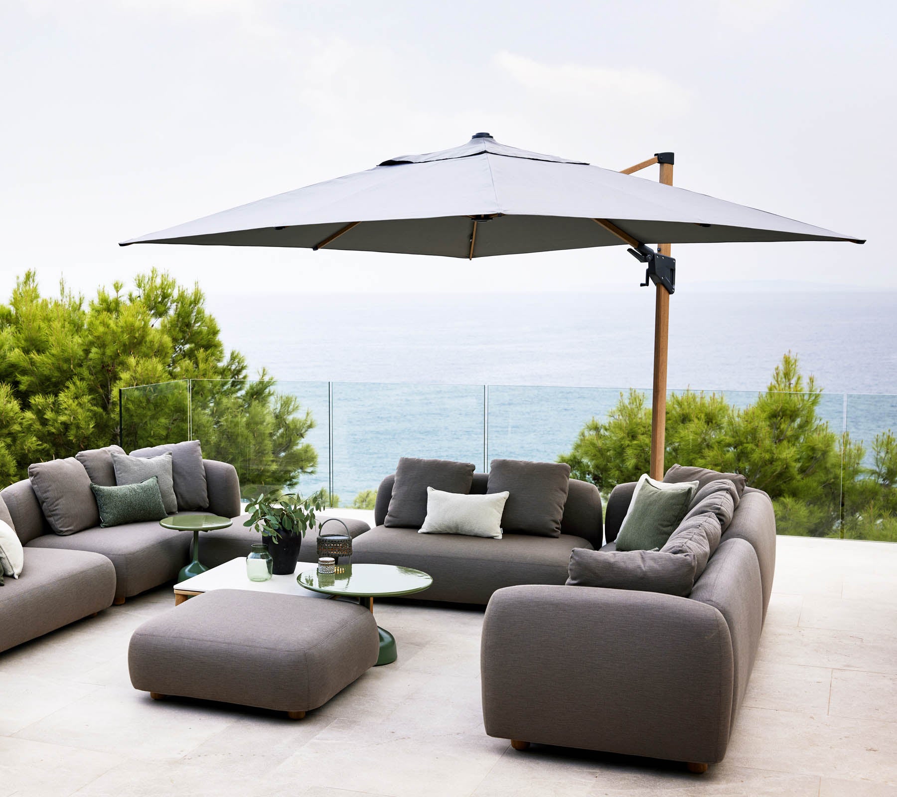 Stylish outdoor seating area with a large umbrella, grey sofas, and a glass table, overlooking a serene water view and lush greenery.
