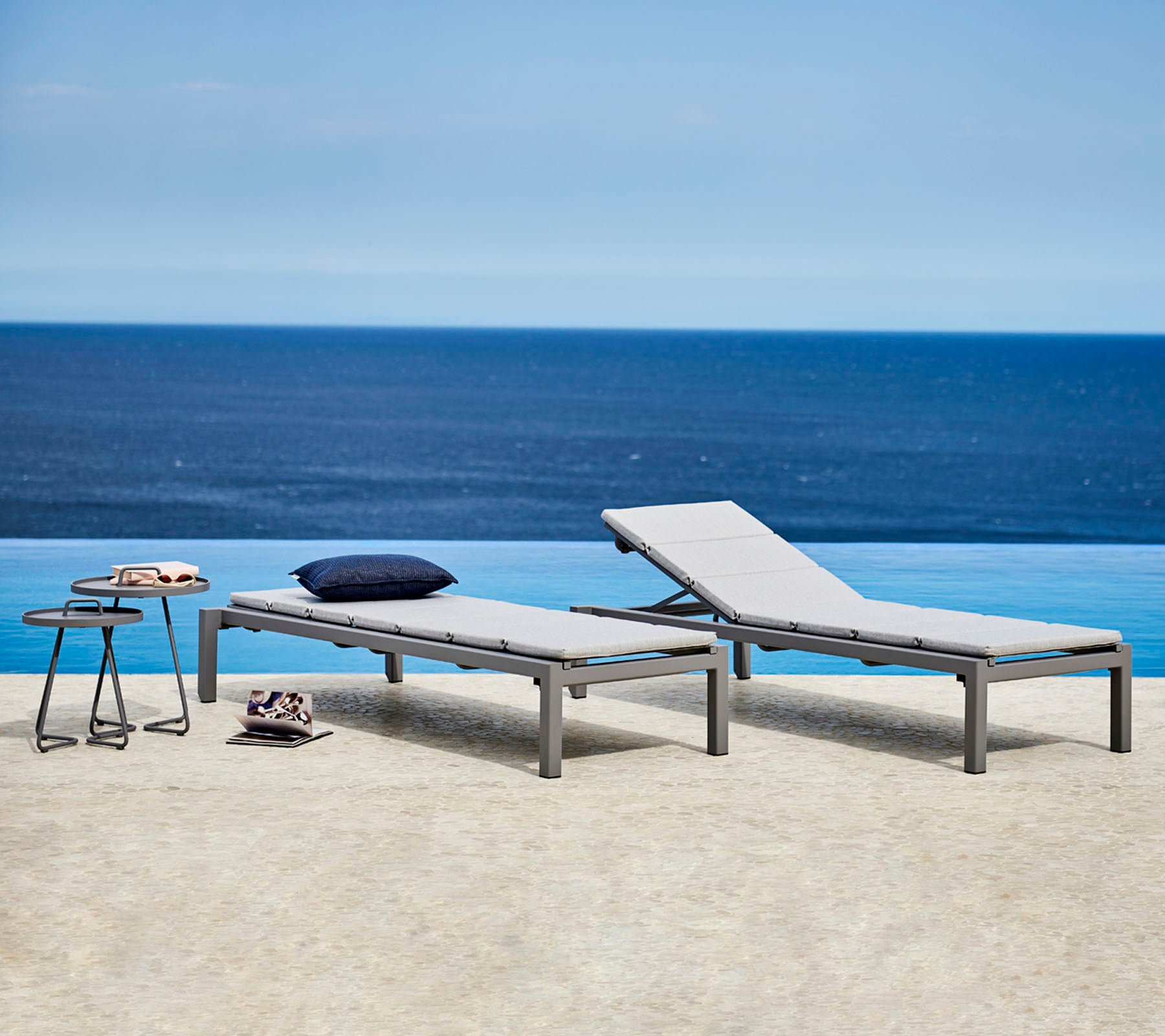 Two stylish lounge chairs with gray cushions are placed beside a small table. The backdrop features a serene ocean view.