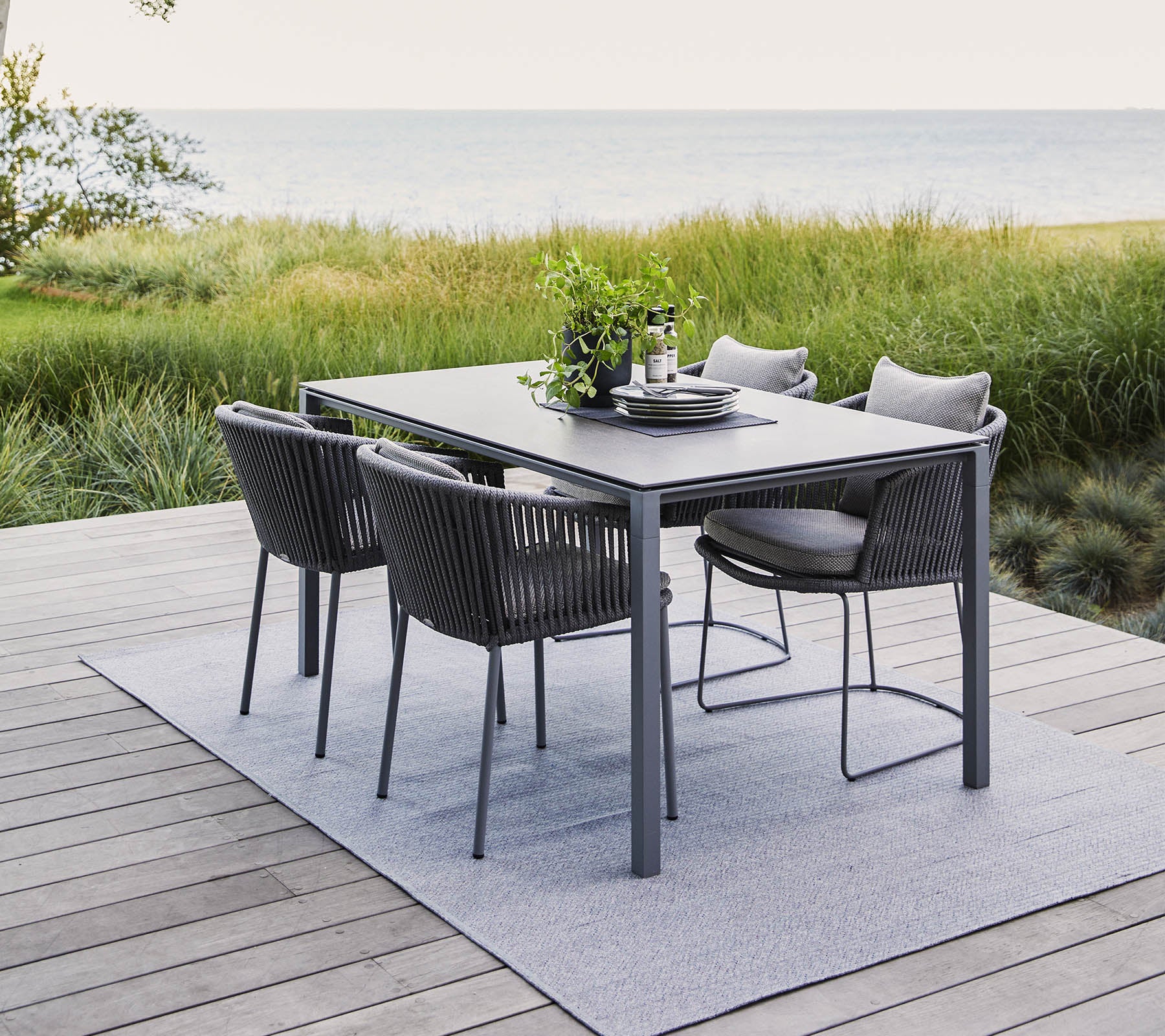 A modern outdoor dining set featuring a rectangular table and four stylish chairs, surrounded by greenery and a serene water view.