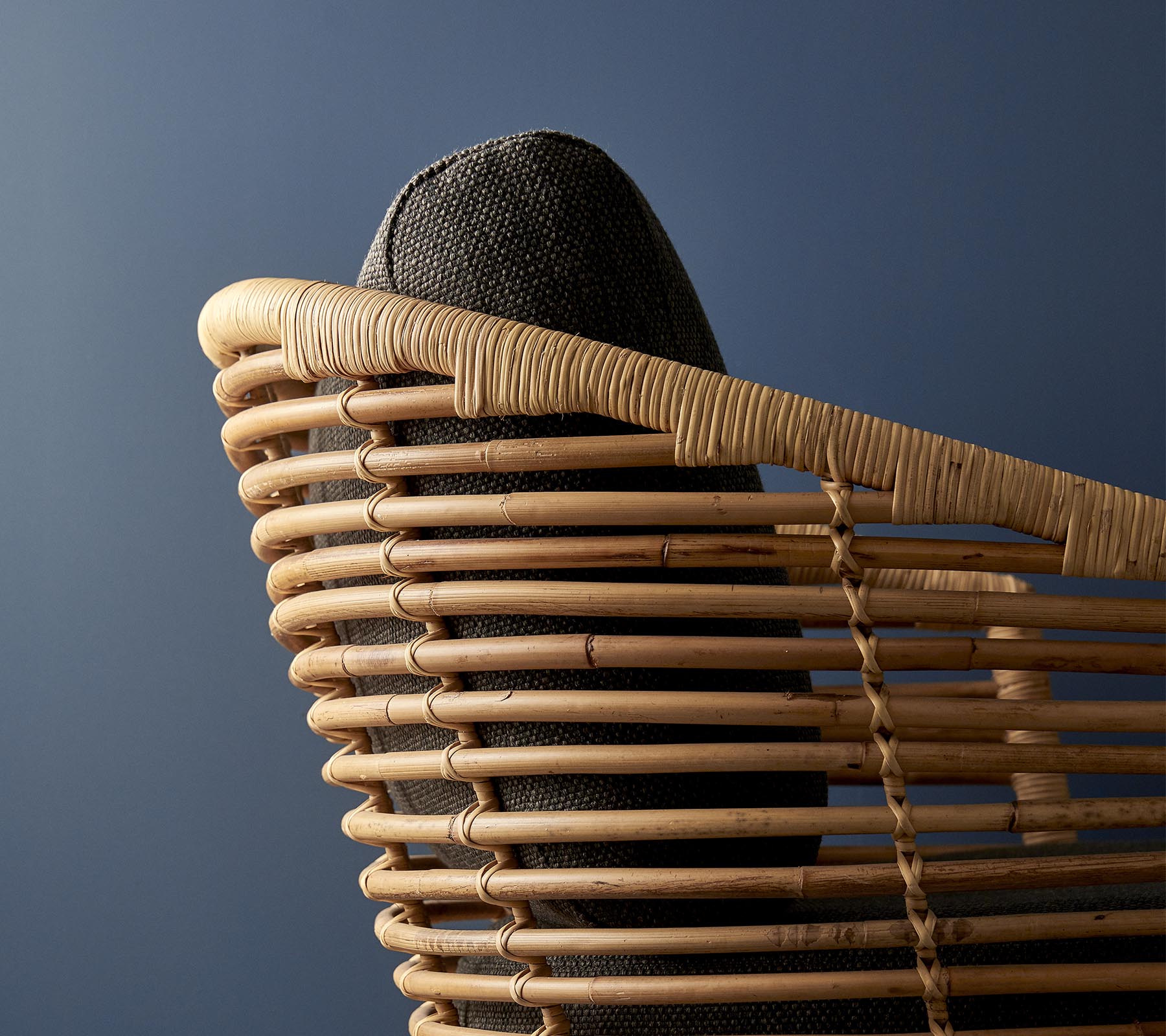 Stylish woven chair with a curved backrest and dark cushioned seat against a muted blue background.
