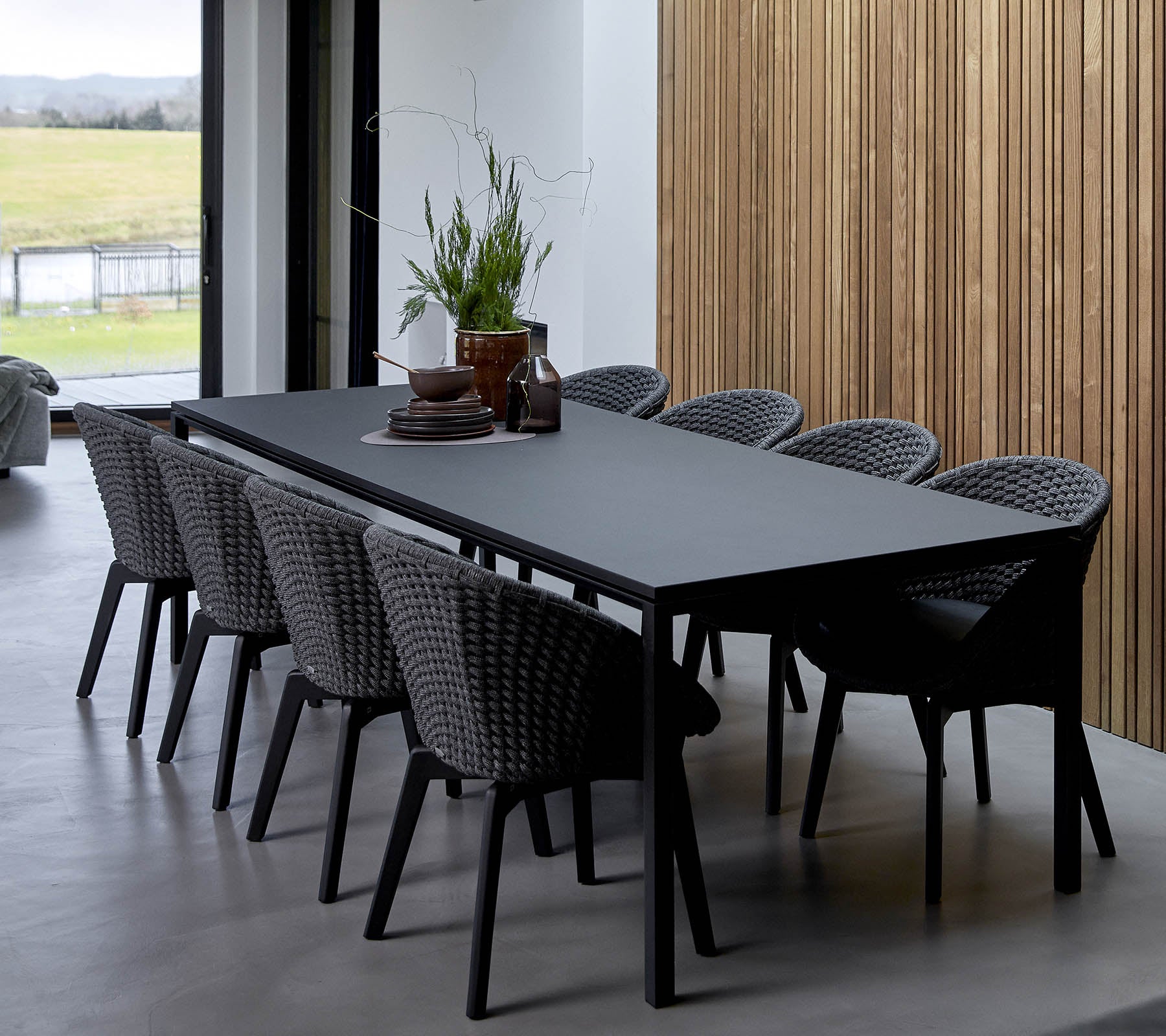 A sleek table surrounded by woven chairs, adorned with decorative plants and a centerpiece, set against a wooden backdrop.