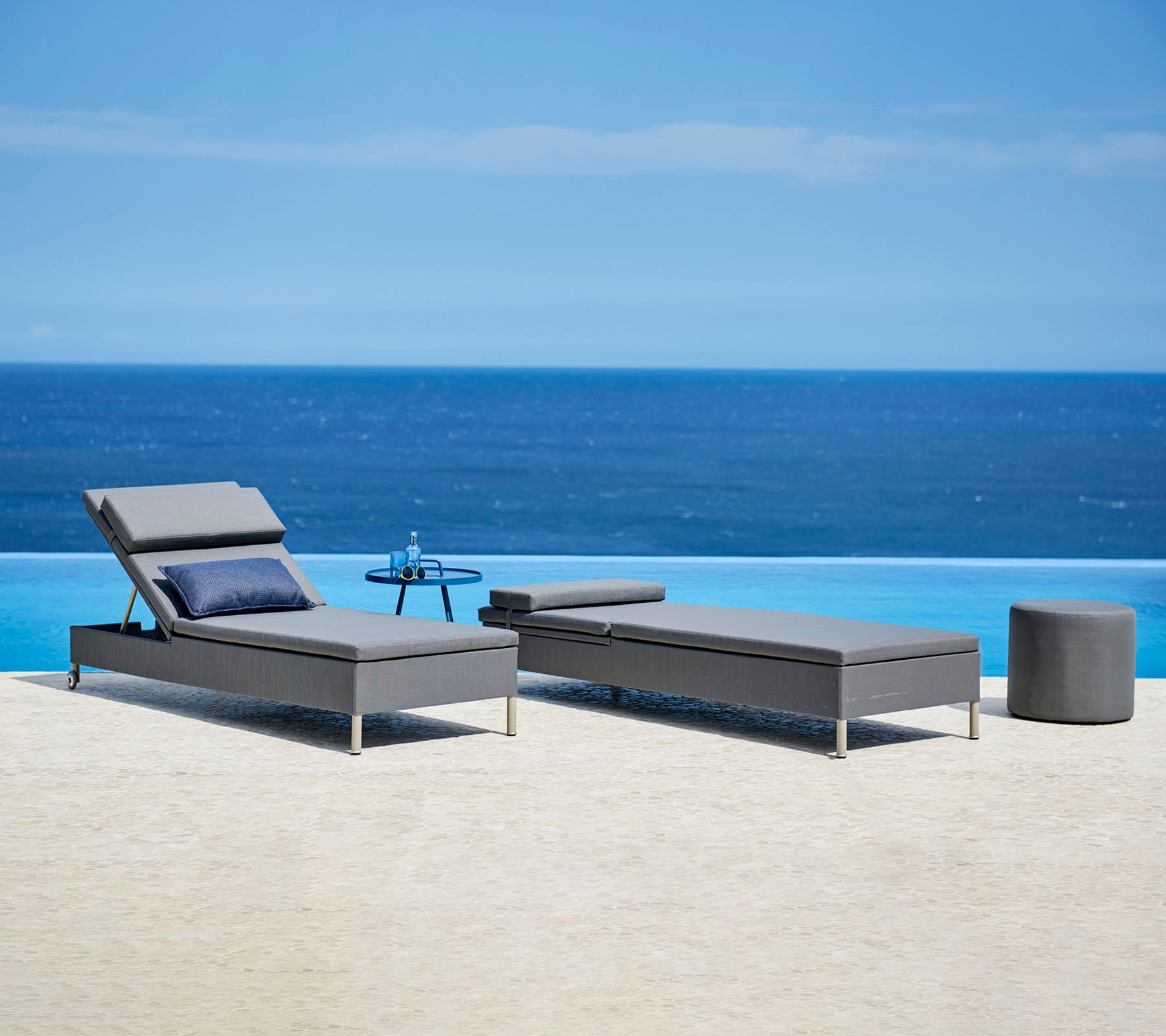Two stylish gray loungers with cushions and a small side table by a serene blue water feature, perfect for relaxation.