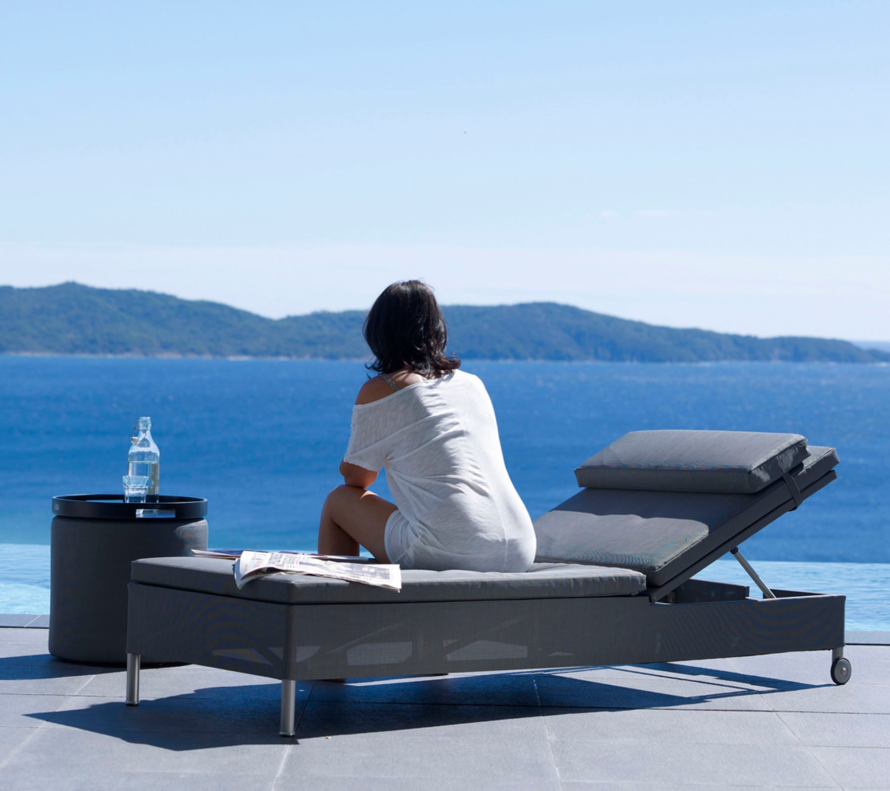 A woman sitting on a lounge chair, looking at a serene ocean view, with a water bottle on a small table beside her.