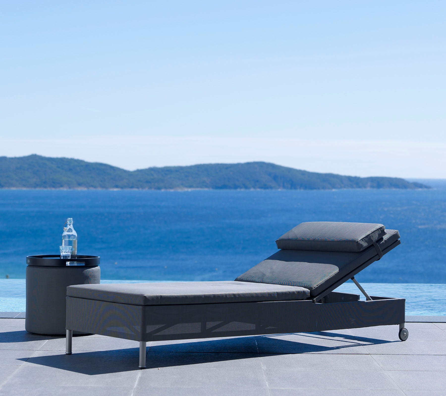 A modern lounge chair and side table beside a serene body of water with distant hills under a clear sky, perfect for relaxation.