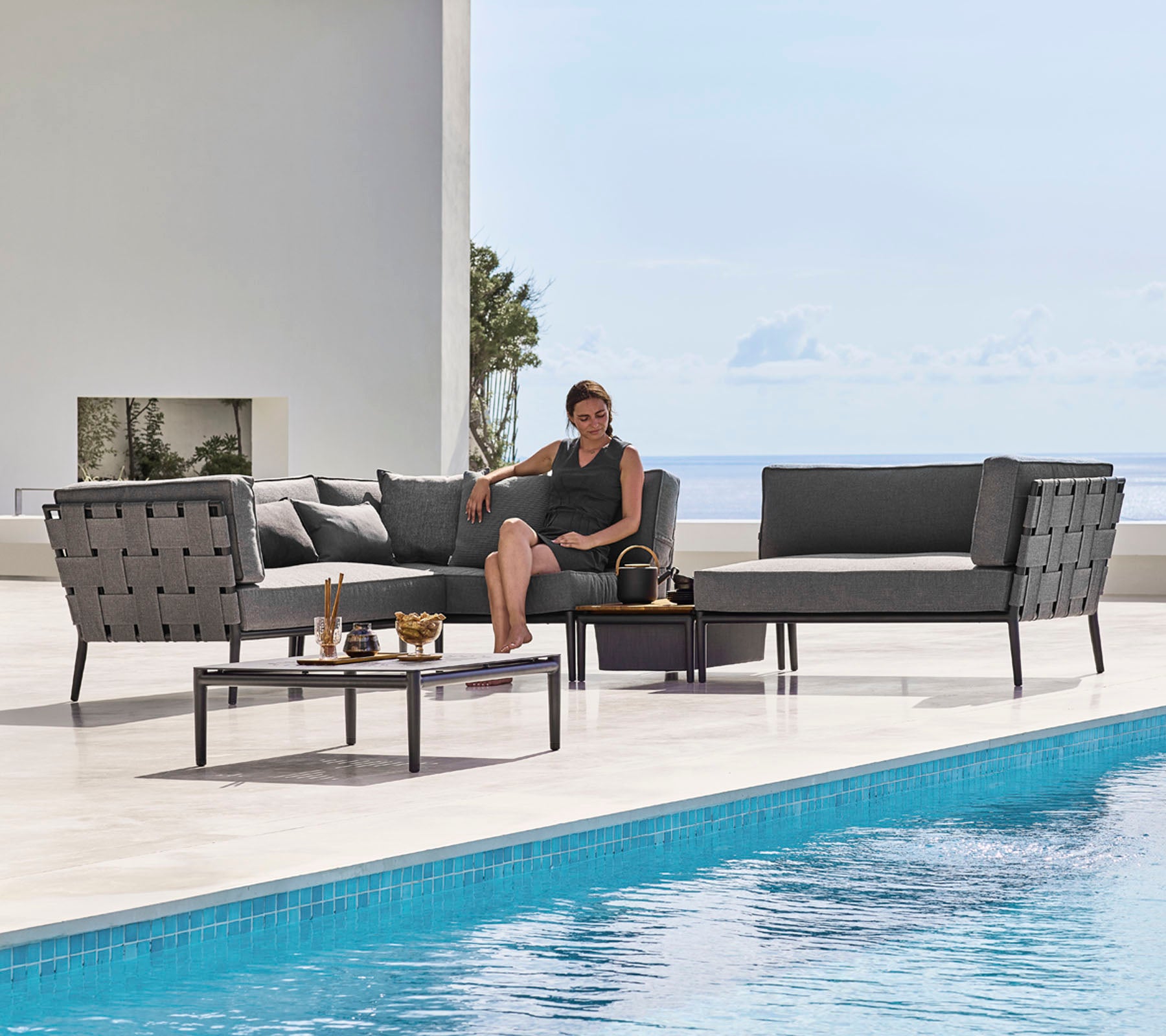 A stylish outdoor lounge set featuring sleek seating and a glass-top coffee table beside a pool, with a person relaxing comfortably.