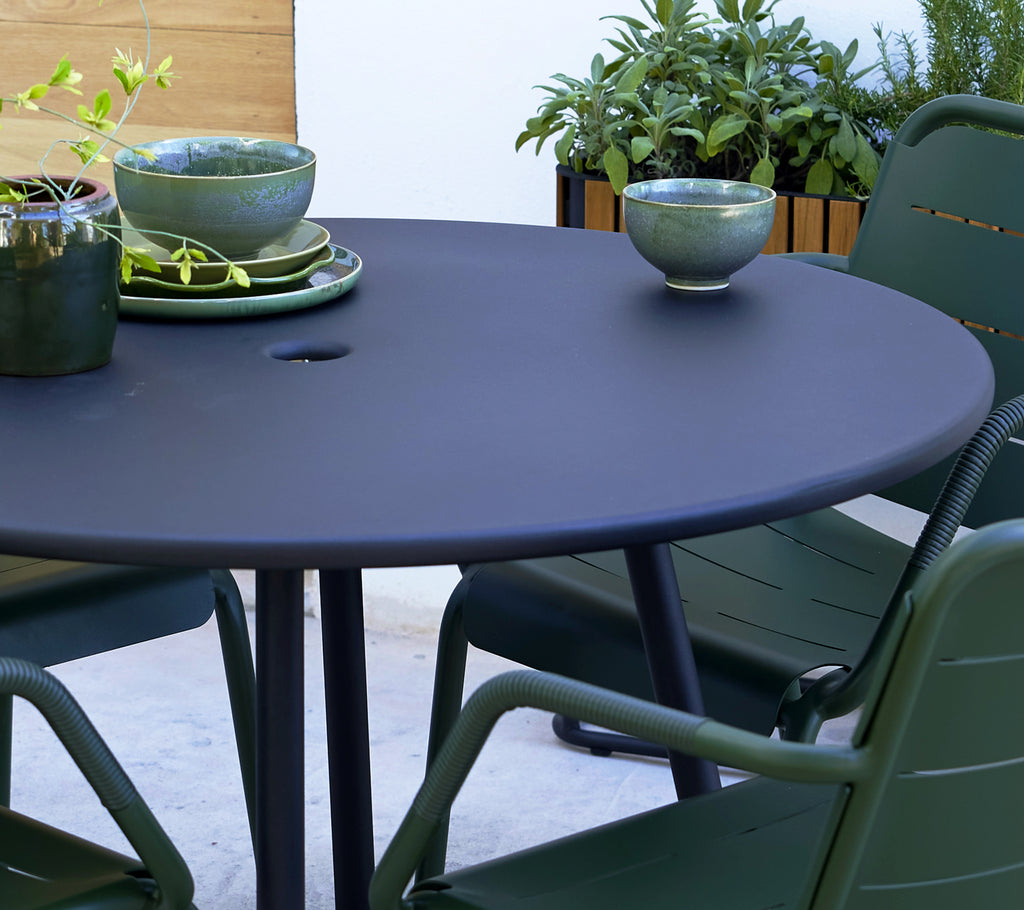 Round grey table with green Copenhagen armchairs, featuring decorative bowls and greenery in the background.