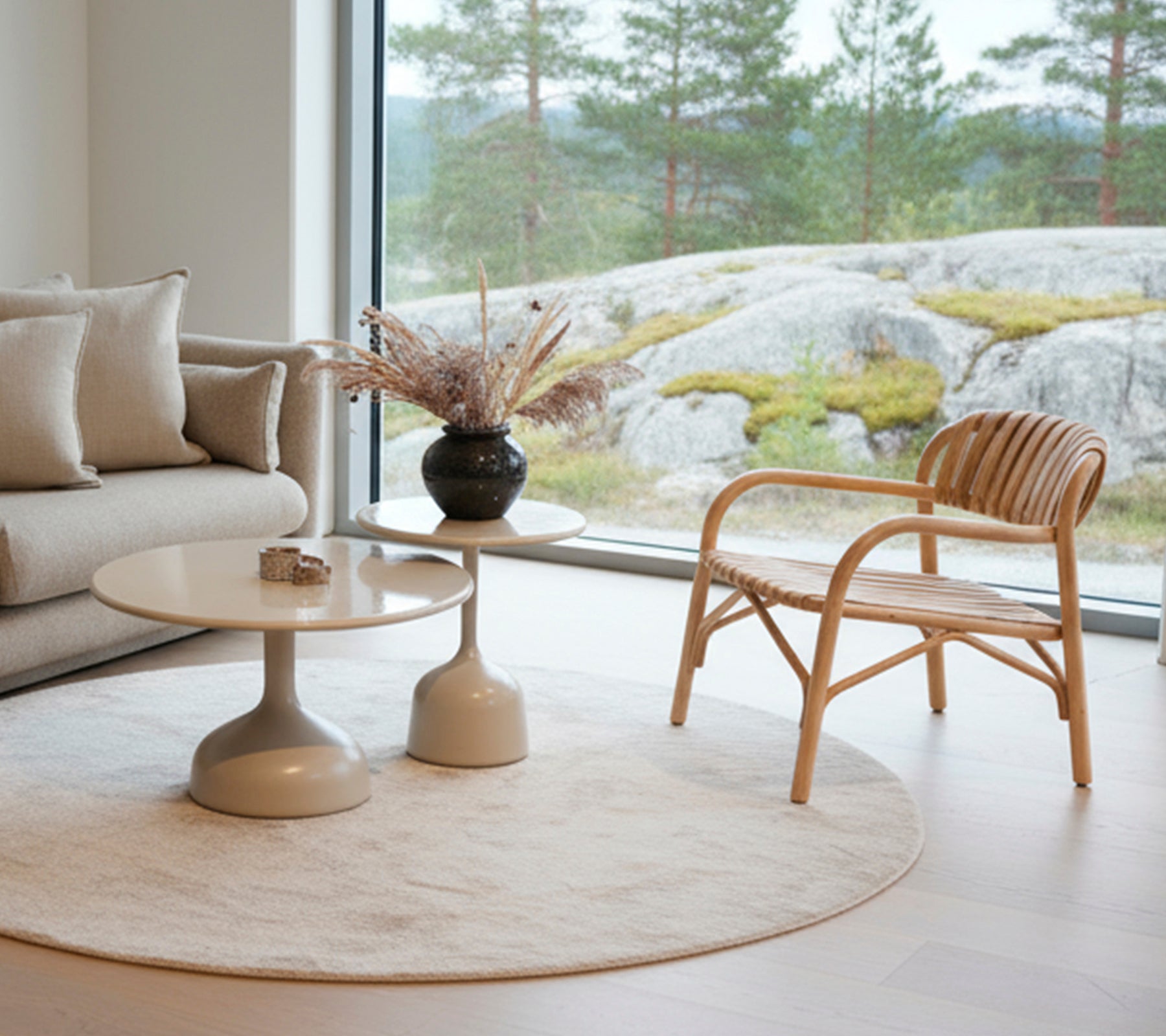 Modern lounge with a sofa, round table, black vase, and rattan chair near a large window.