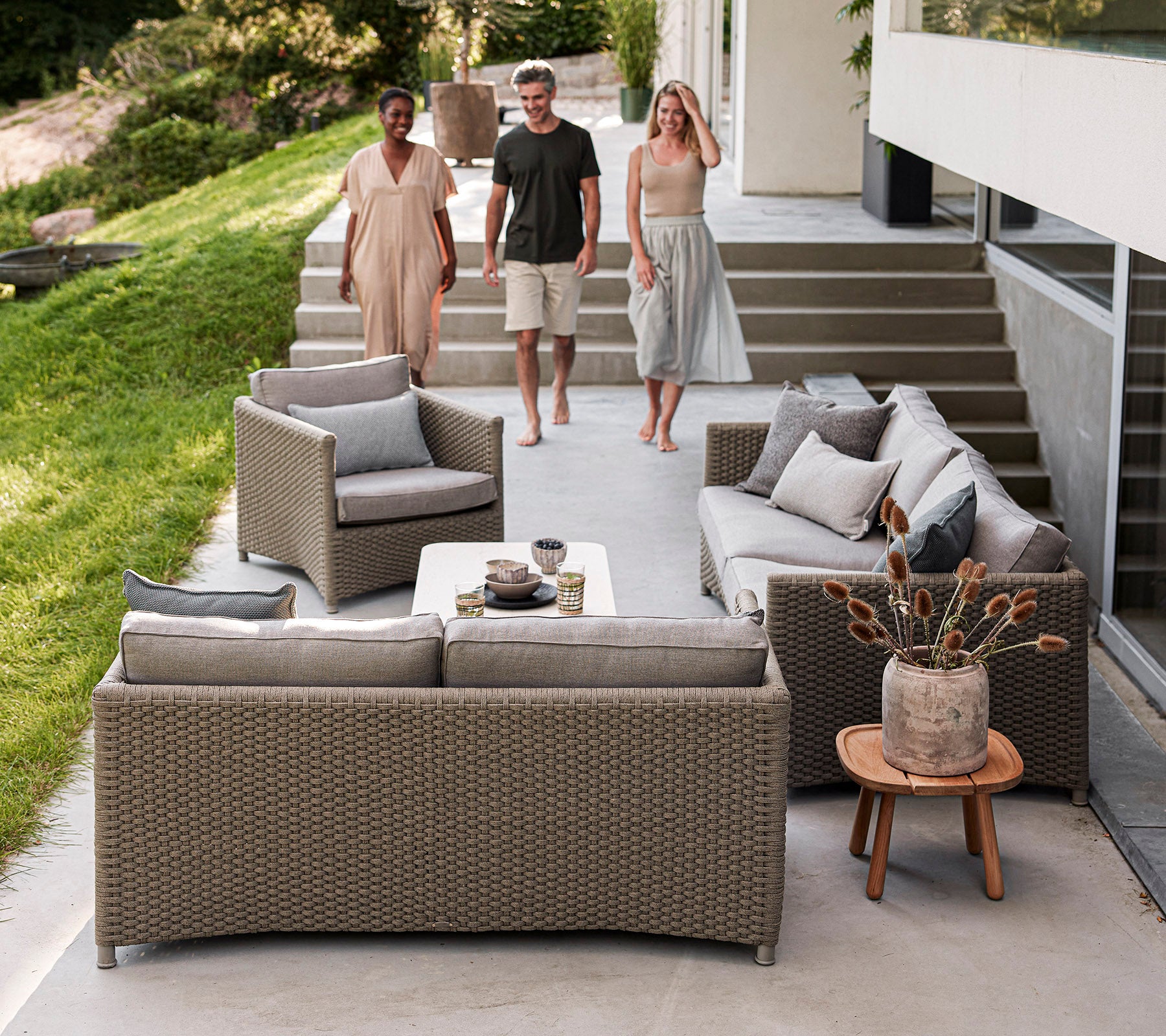 Three people walk toward a stylish outdoor seating area featuring cozy, woven furniture with decorative pillows and a coffee table.