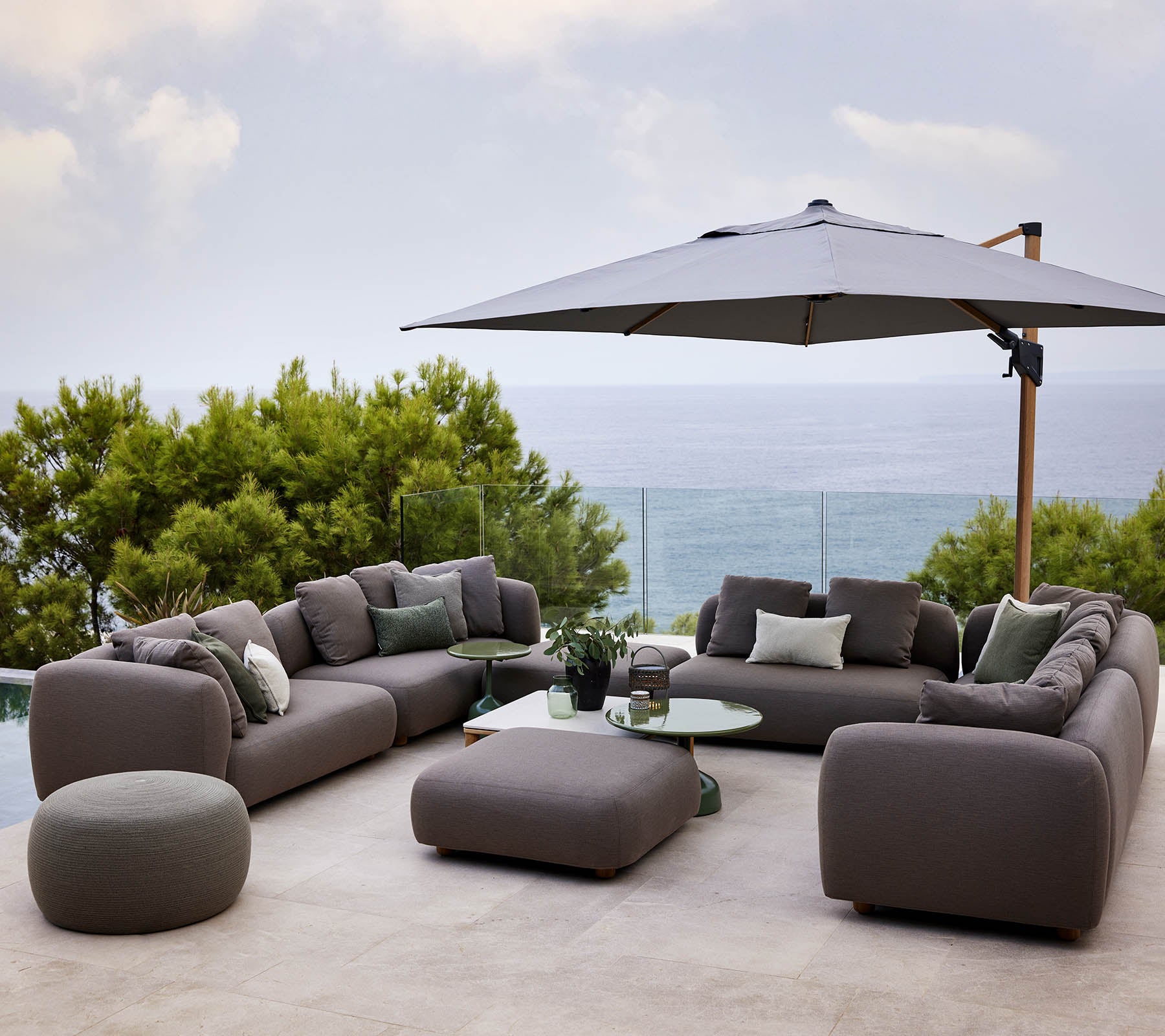 Modern lounge seating set with plush cushions, accompanied by a round table and a large umbrella, surrounded by greenery and a scenic view.