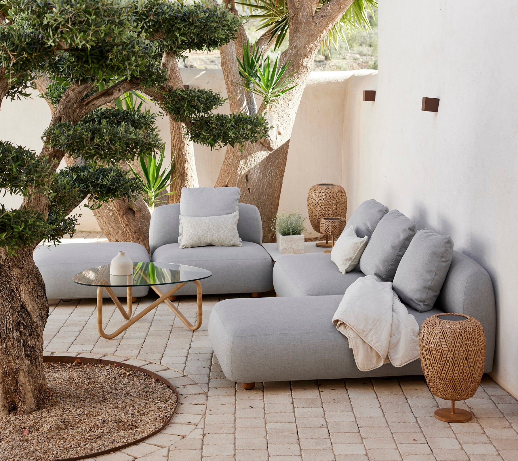 Modern lounge area featuring a grey sofa, cushions, glass coffee table, and woven side table.