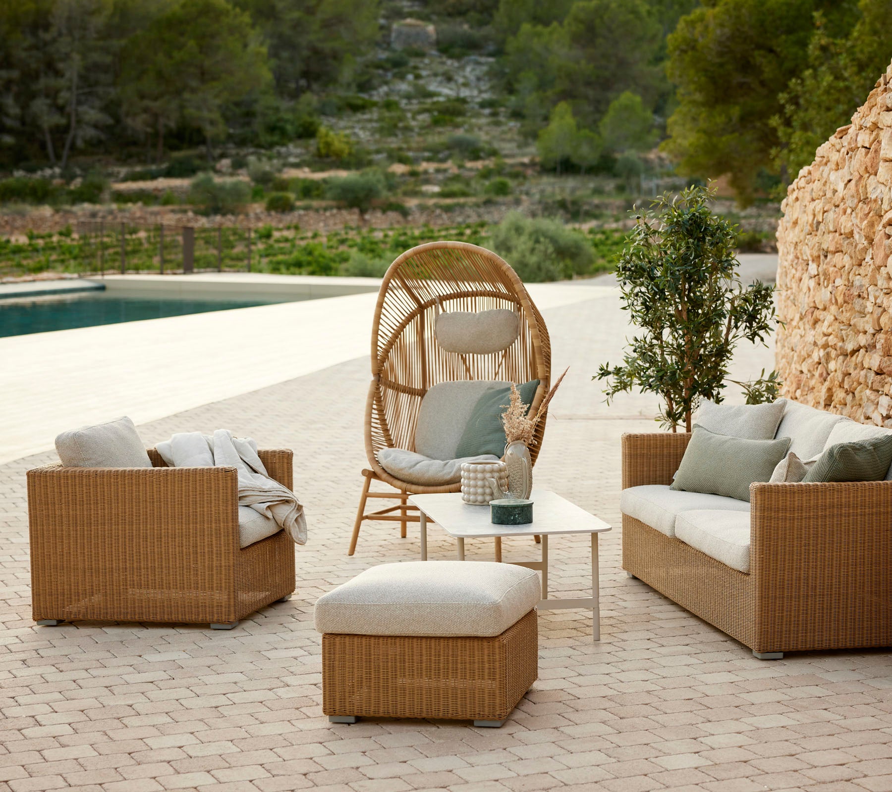 Cozy outdoor seating arrangement featuring woven armchairs, a circular hanging chair, an ottoman, and a small table among greenery.