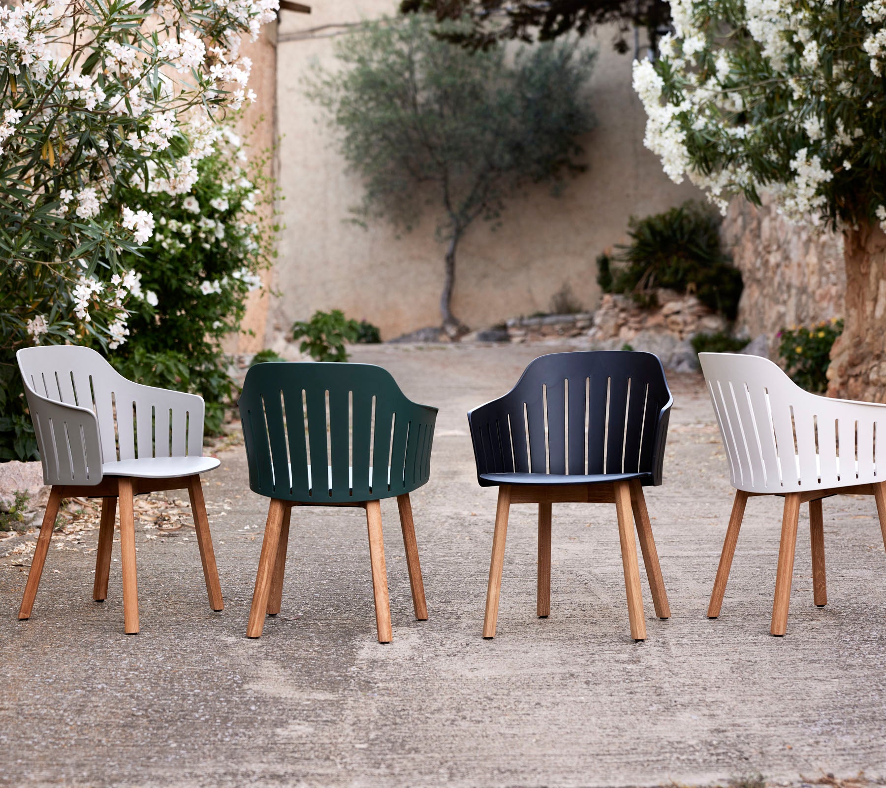Four stylish chairs in different colors with wooden legs, showcasing a modern design and slatted backrests amidst greenery.