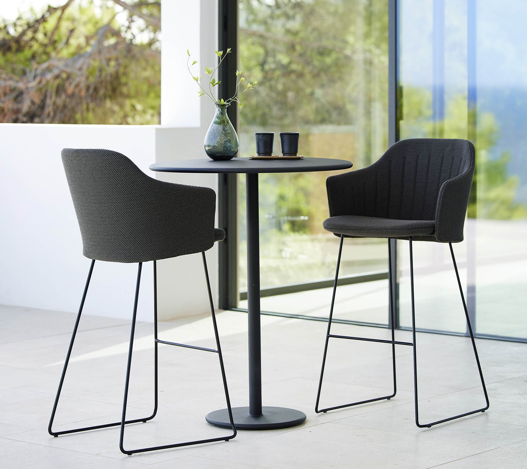 Two stylish choice bar chairs paired with a sleek round bar table, accented by decorative items and a natural backdrop.