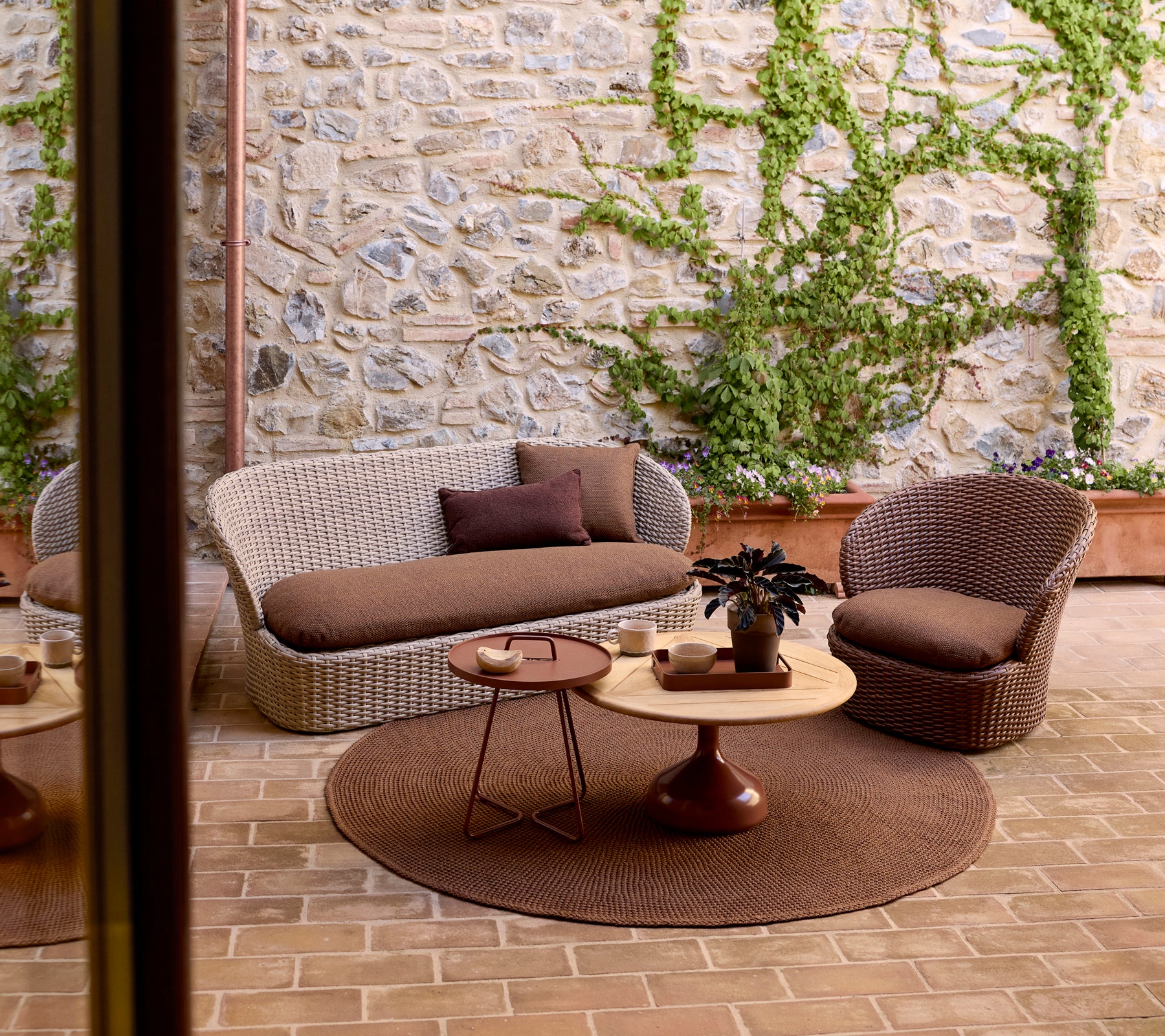 Cozy outdoor lounge area featuring a woven sofa, a chair, a round side table, and greenery against a stone wall.