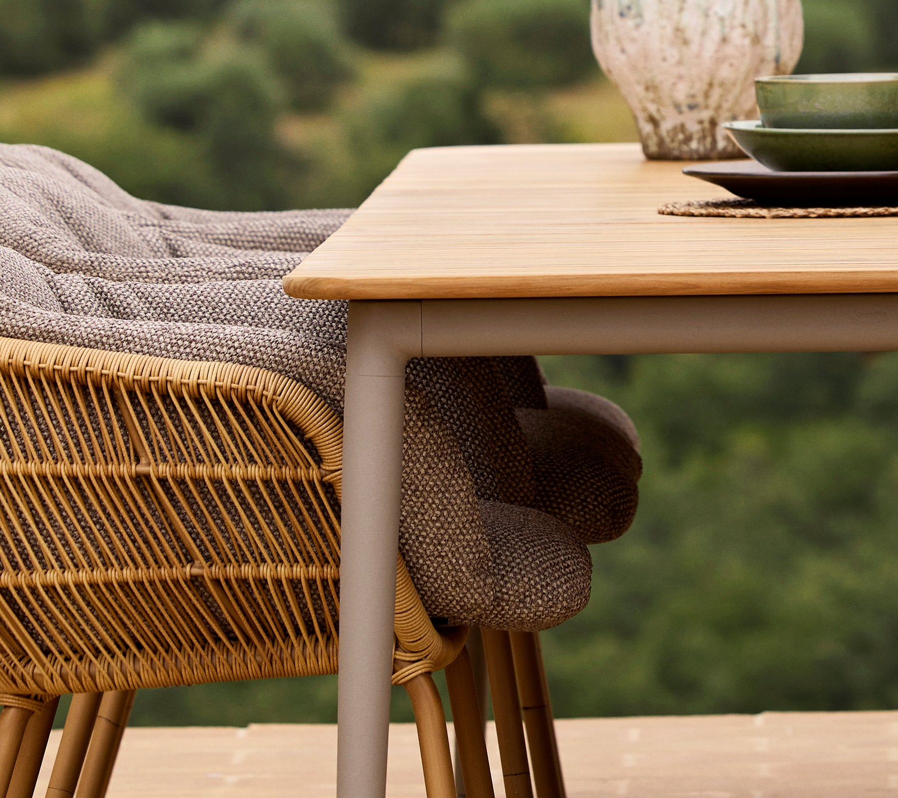 A close-up of a Core dining table with a Stay chair, featuring woven fabric and a green plate set against a nature backdrop.