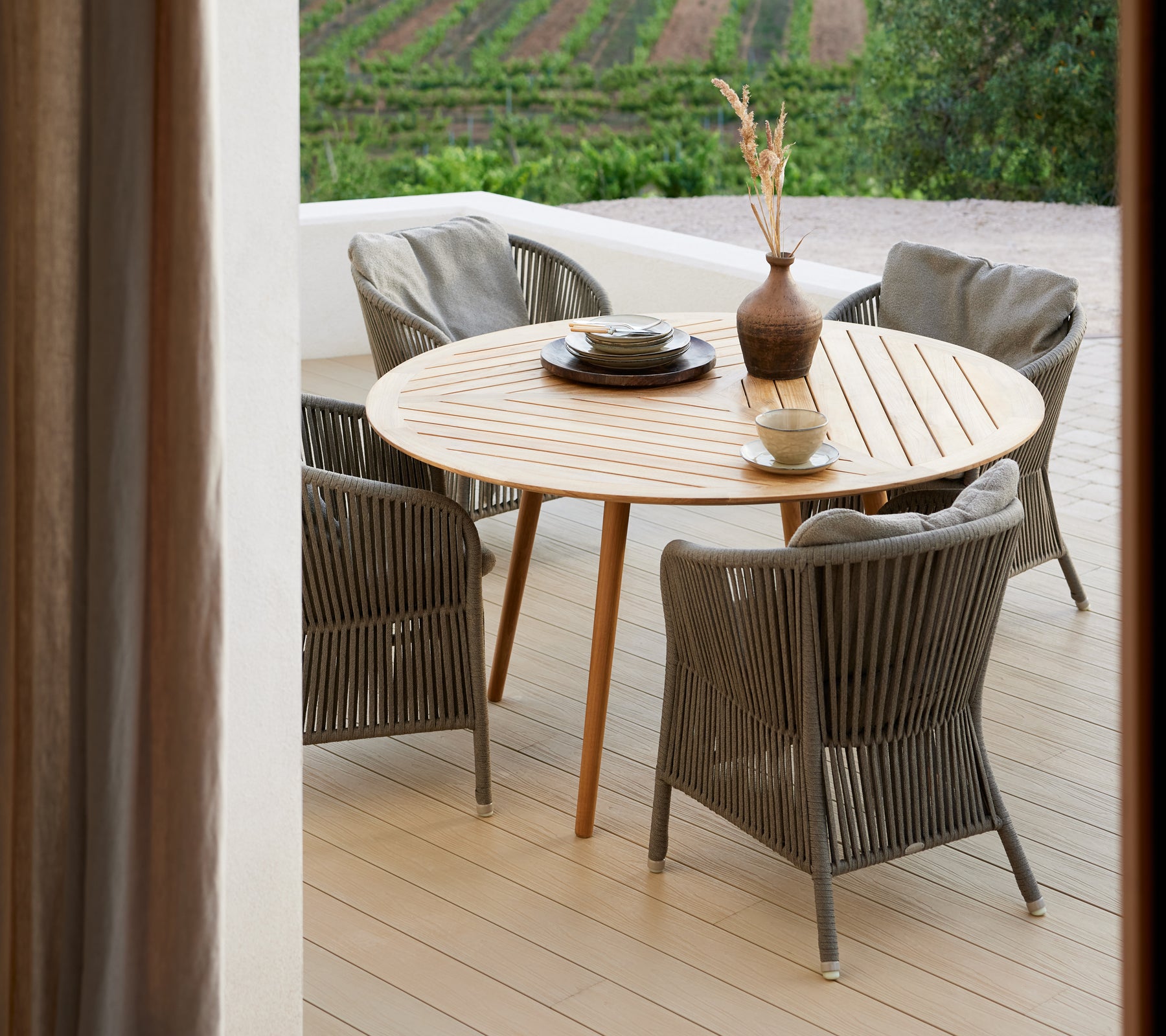 A round wooden table surrounded by four woven chairs, featuring a vase and decorative dish, set against a scenic backdrop.