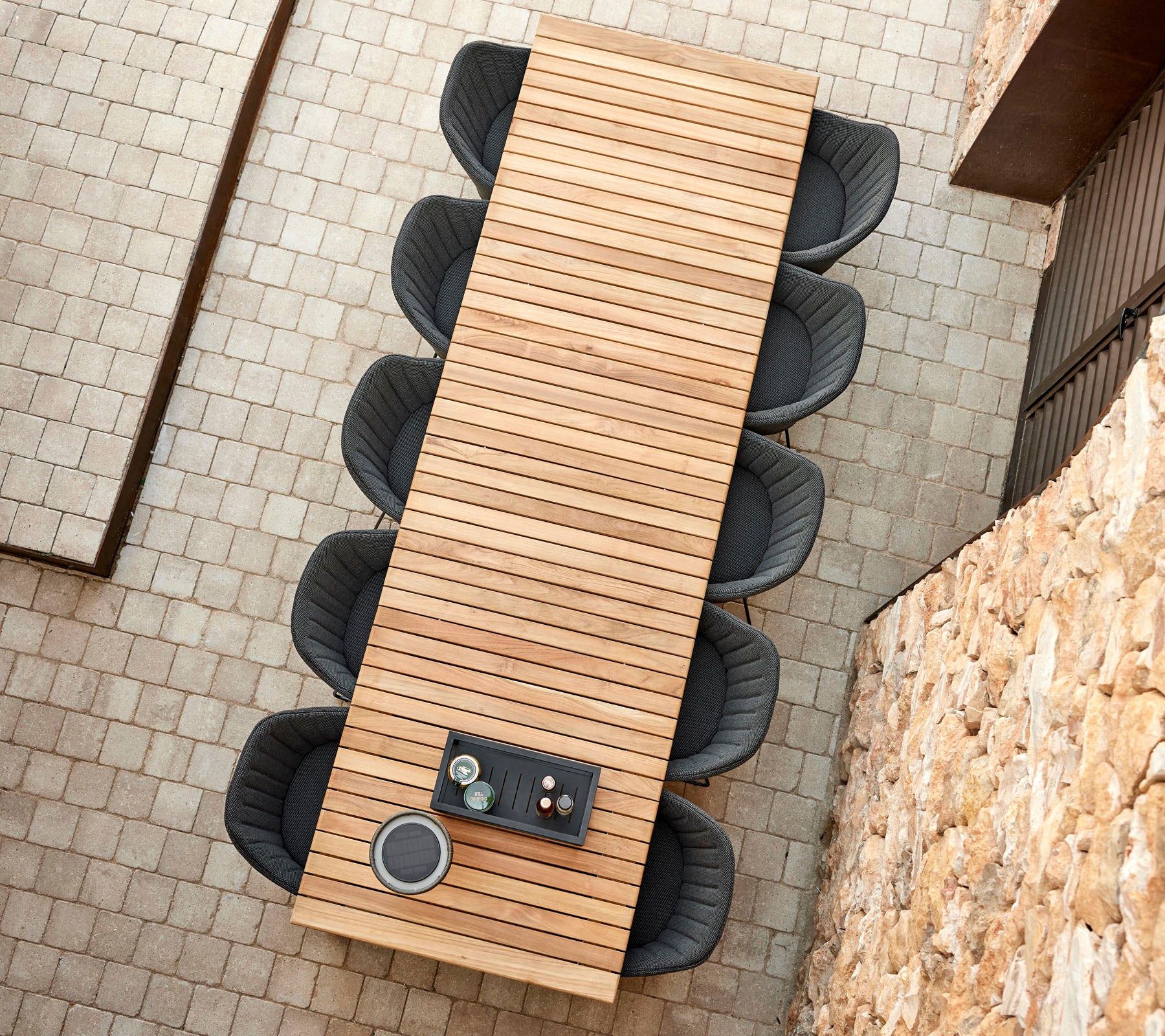 A long wooden table with slatted design, surrounded by circular black chairs, featuring a small tray with cups on top.