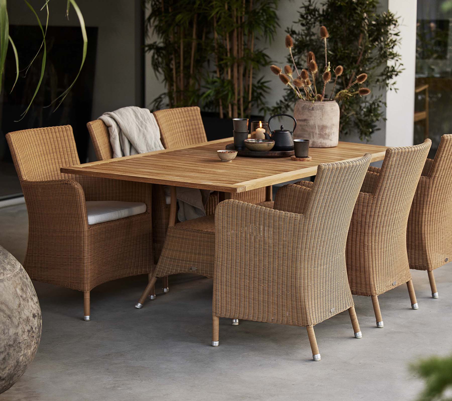 A dining setup featuring a rustic wooden table surrounded by woven Hampsted chairs, complemented by decorative plants.